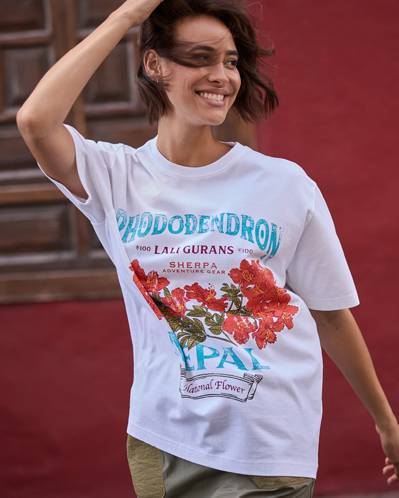 A woman wearing a white t-shirt with floral design and the text "Rhododendron" printed on the front. The woman is turned to the side as she looks over her shoulder with the wind blowing her hair. She is standing outside, against a red plastered wall with a brown wooden window.