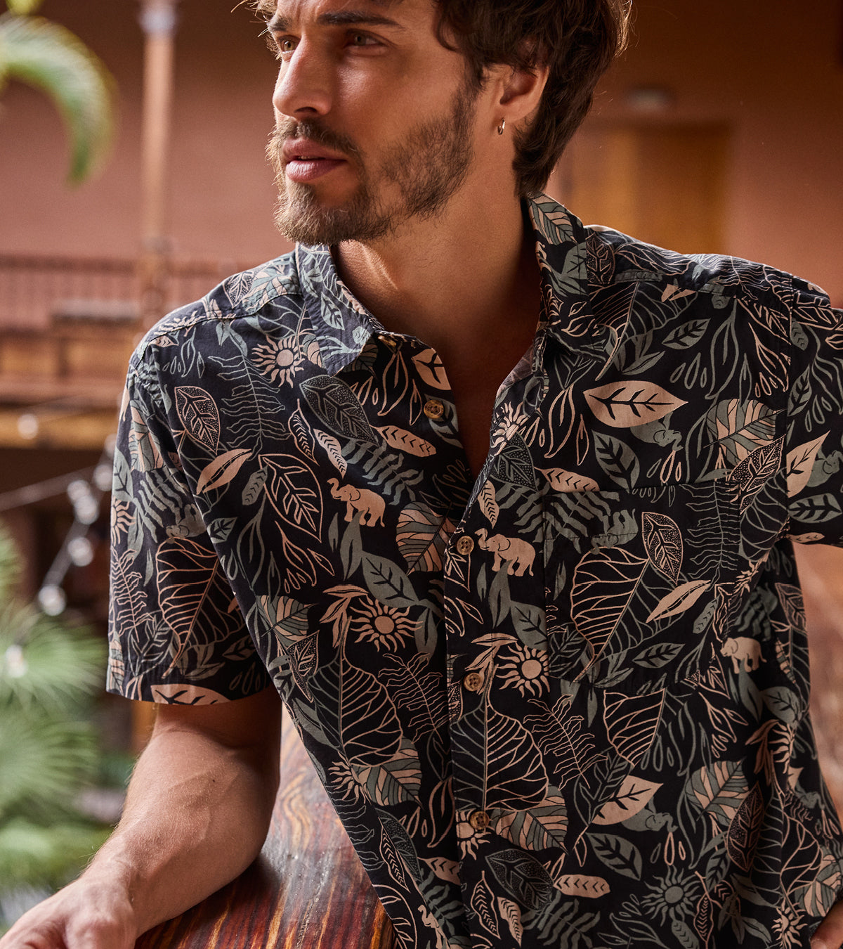 Man wearing a black patterned shirt sitting at a table with a blurred background. The patterned shirt depicts green and cream leaves and elephants.