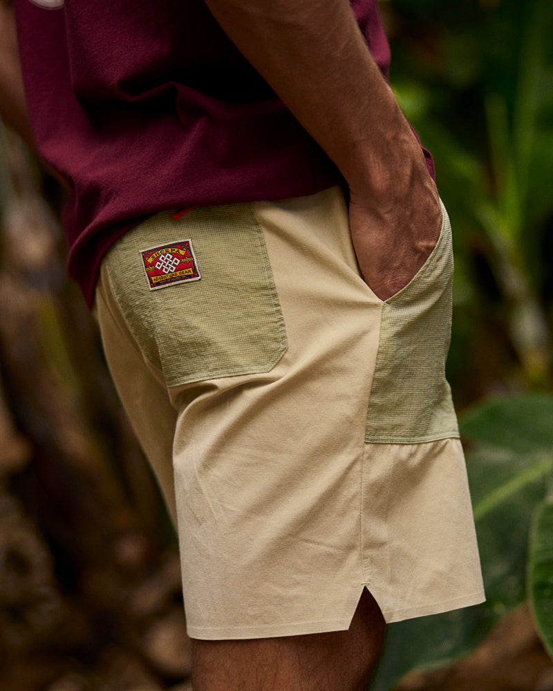 A close up of a man wearing a pair of beige shorts with a patch on the pocket and a burgundy T-Shirt.