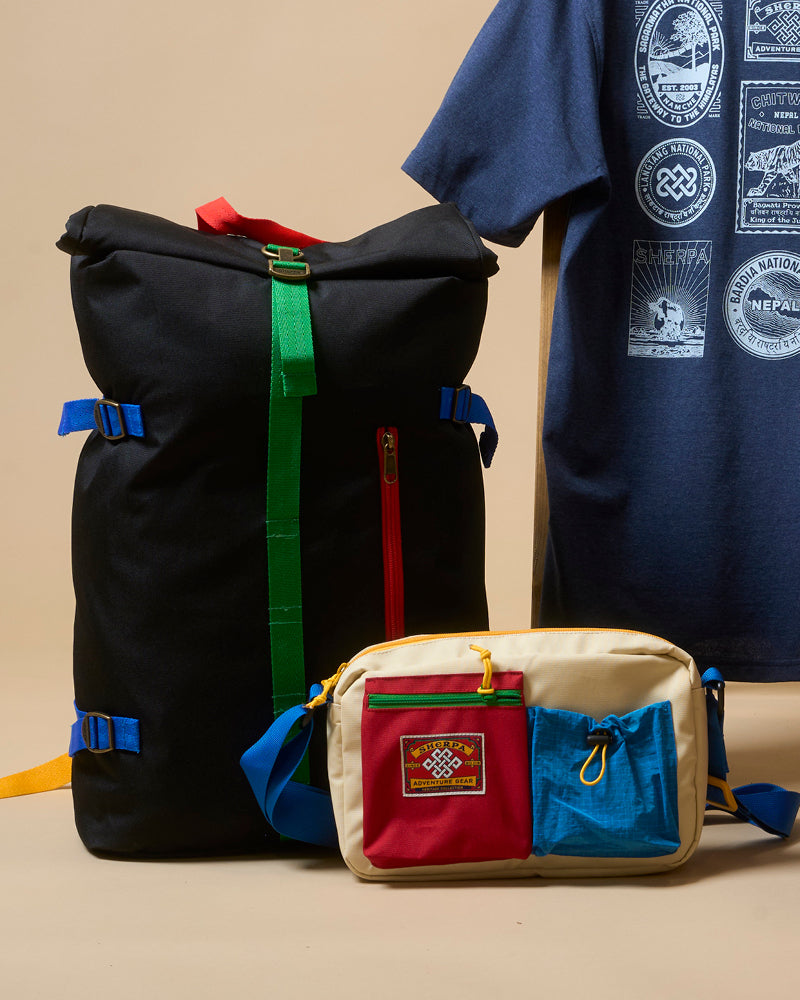 Black backpack with colorful straps next to a navy t-shirt with graphic designs on a beige background. In front of the backpack is a smaller, beige hip bag with colorful pockets and straps. A small patch is stitched onto the front of one of the pockets, depicting a Nepali endless knot and the words "Sherpa Adventure Gear".