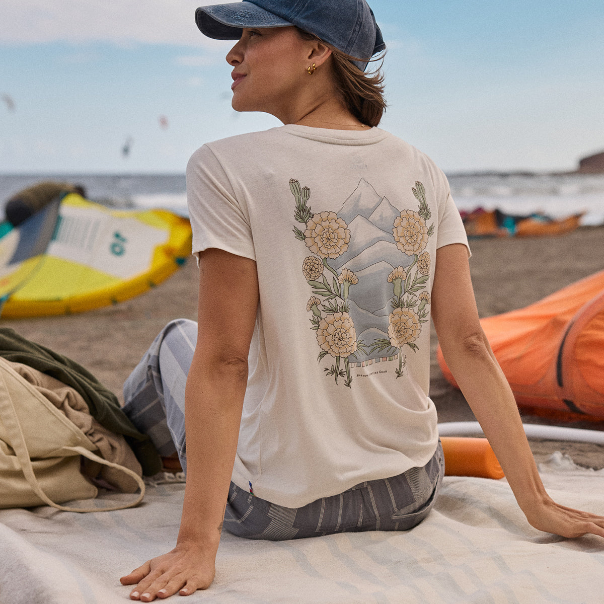 A woman sitting on a beach wearing a t-shirt with a nature-themed design, surrounded by beach gear. The T-Shirt depicts a blue mountain scene with cream and yellow flowers.