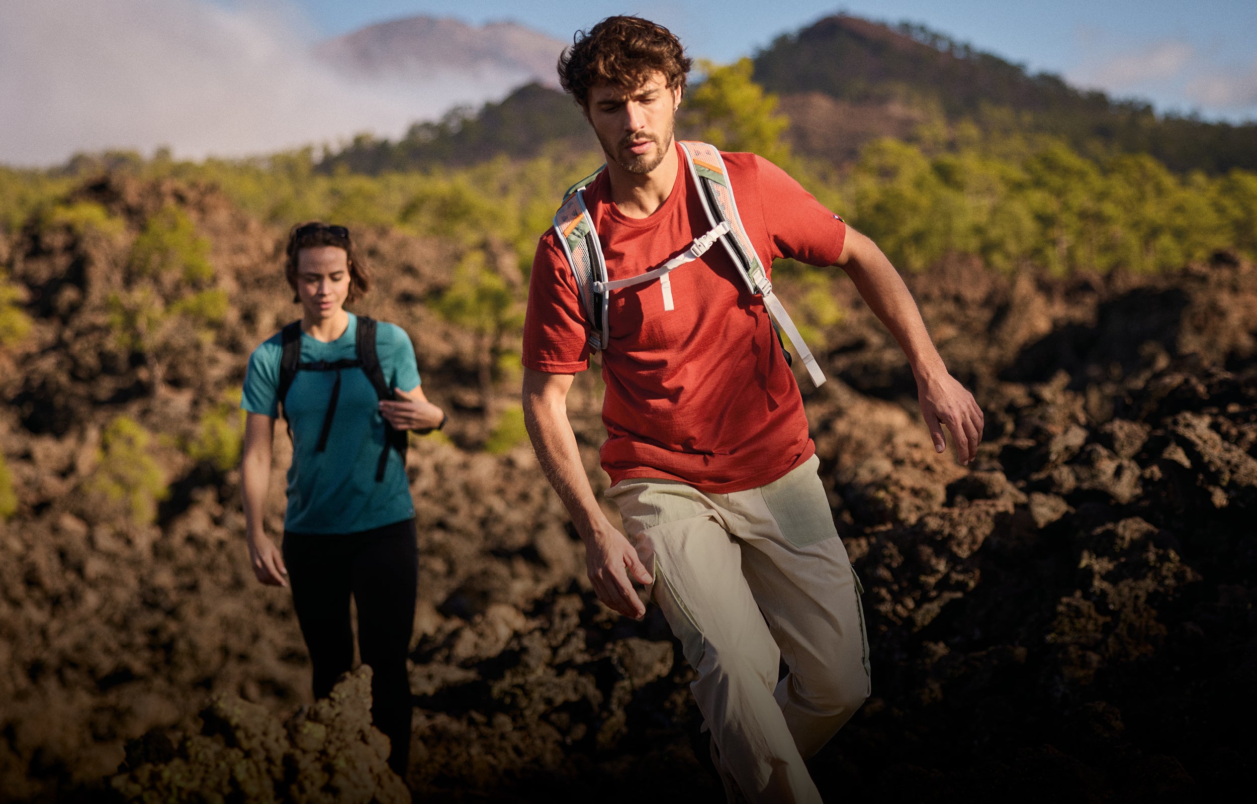 A man and a woman on a trail with mountains in the background. The main us wearing a red T-Shirts and a pair of cream cargo trousers with green pockets and a green bag. The woman is wearing a teal T-Shirt, a pair of black leggings and a black backpack.