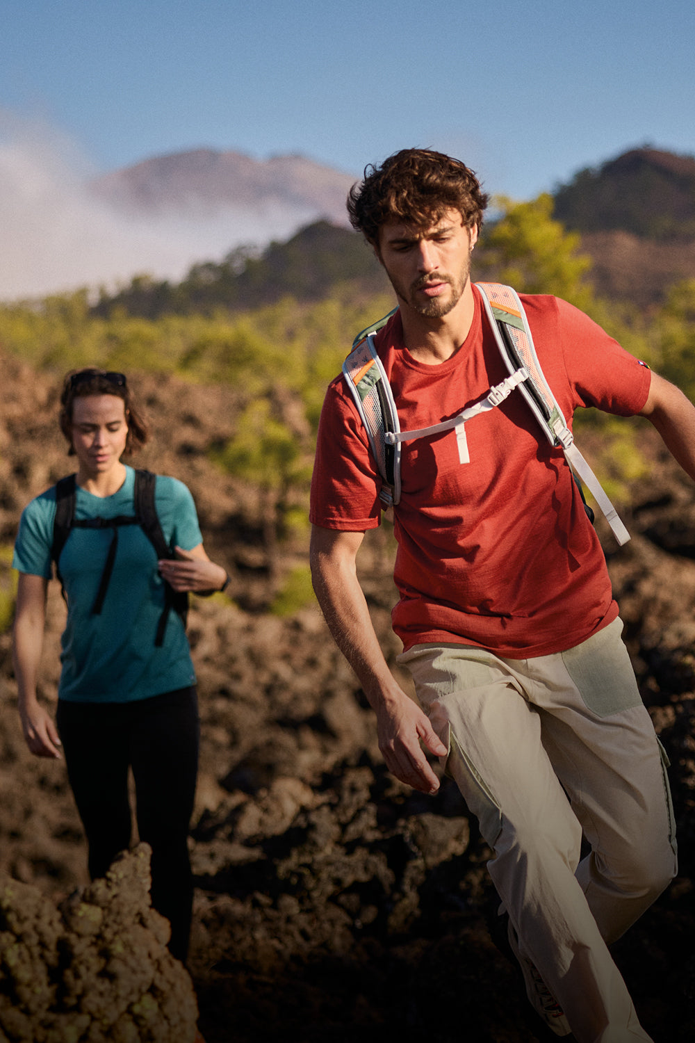 A man and a woman on a trail with mountains in the background. The main us wearing a red T-Shirts and a pair of cream cargo trousers with green pockets and a green bag. The woman is wearing a teal T-Shirt, a pair of black leggings and a black backpack.