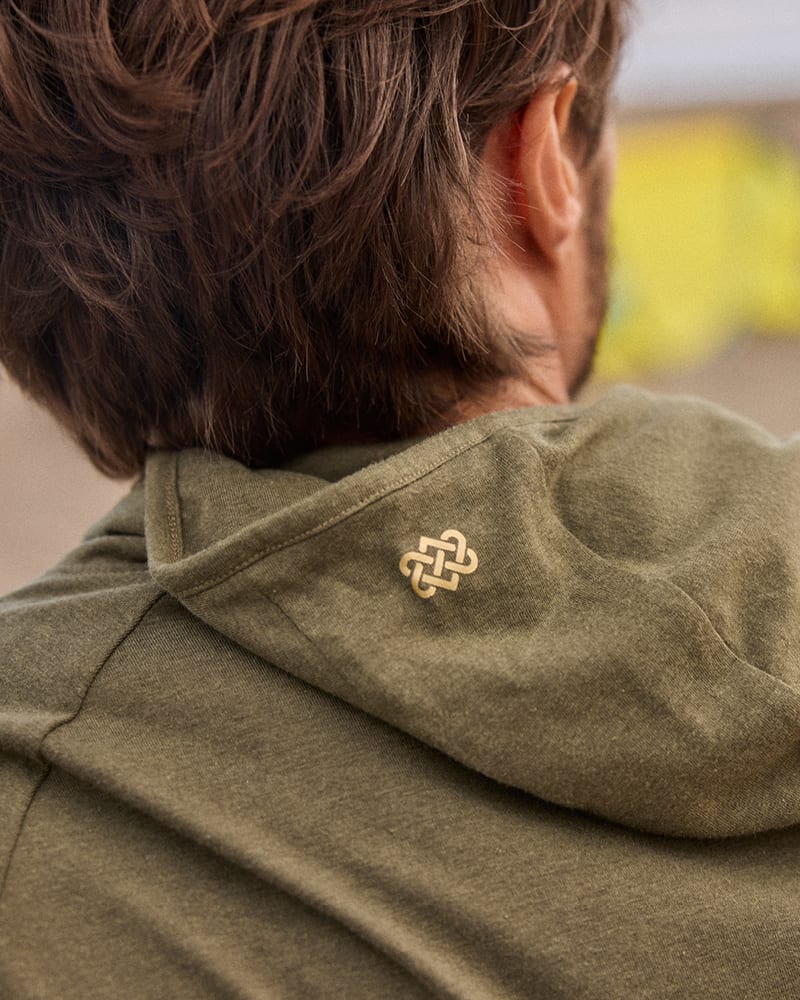 Close-up back view of a person wearing an olive green Sherpa Adventure Gear hooded sweatshirt, showing a small gold-toned endless knot logo on the hood.