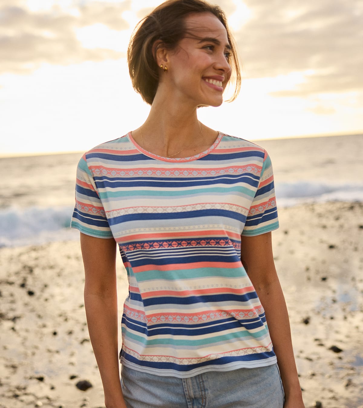 A woman wearing a short-sleeve crewneck t-shirt with horizontal stripes in navy blue, teal, coral pink, and white, standing on a beach at sunset.