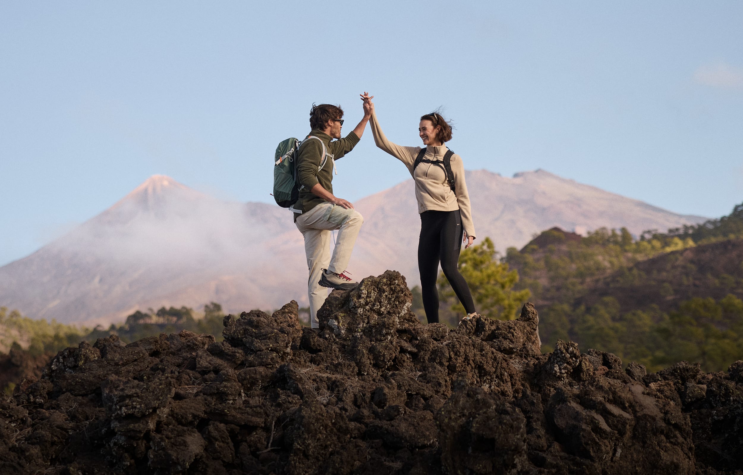 Two hikers high-five on a dark volcanic rock with mountains in the background under a clear blue sky. The man is wearing a green fleece, beige trousers and a green backpack. The woman is wearing black leggings, a beige fleece and a black backpack