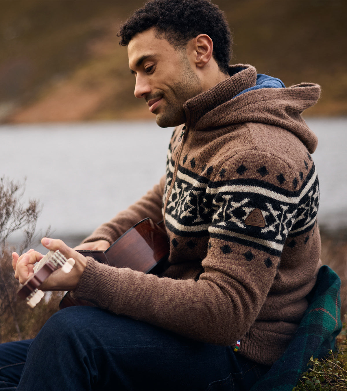 Man in a brown Fair Isle knit hoodie holding a guitar, sitting by the water.
