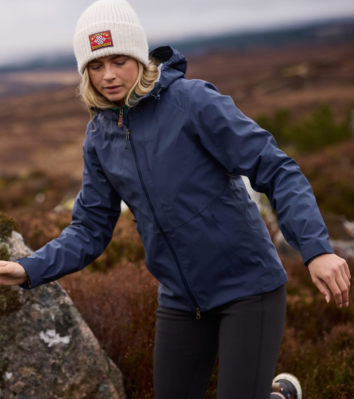Woman hiking outdoors in a navy hooded jacket, black leggings and white knit beanie.