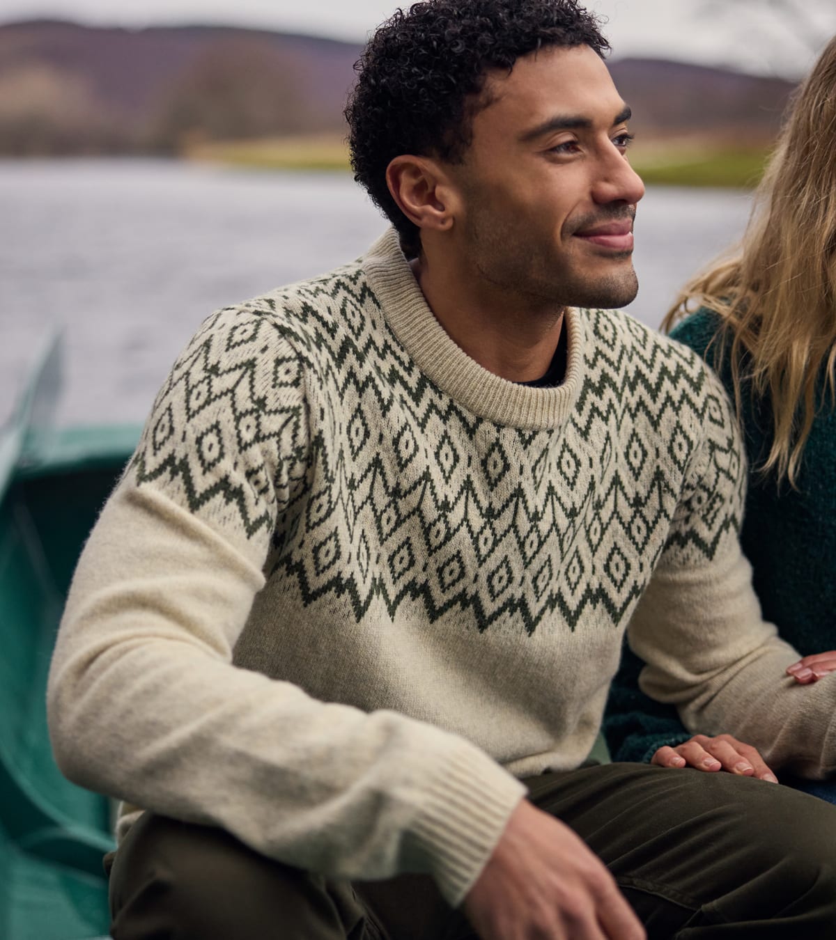 Man in a cream knit sweater with a dark geometric diamond pattern, sitting by a lake.