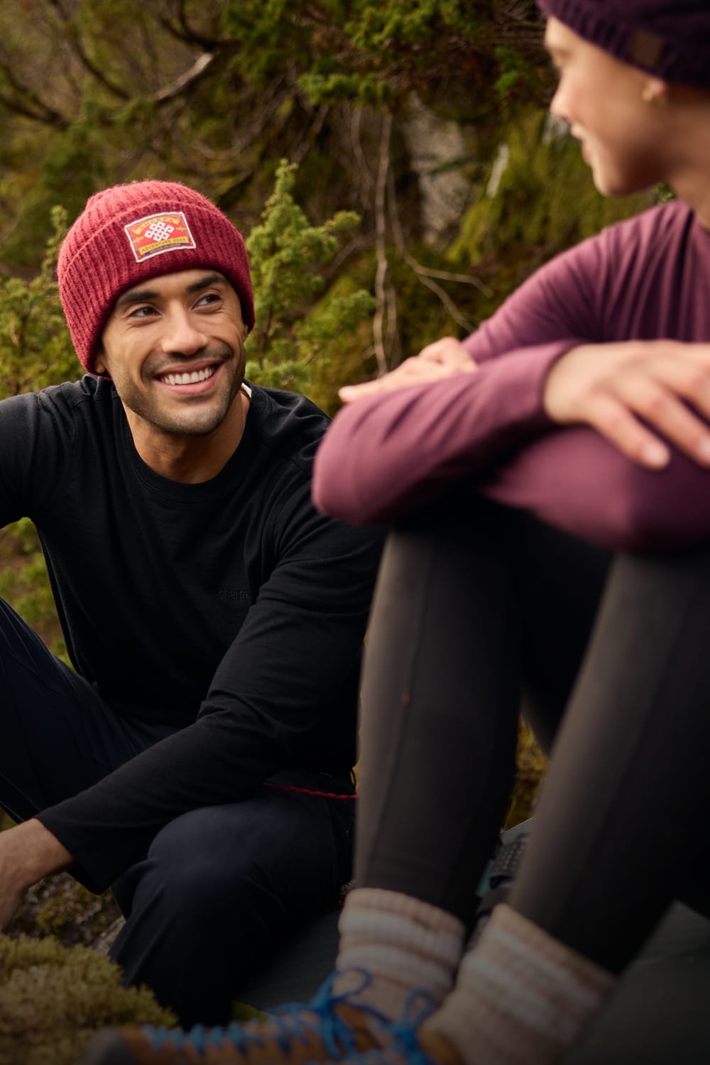 Man wearing a red knit beanie with a visible logo, sitting outdoors in a forest.