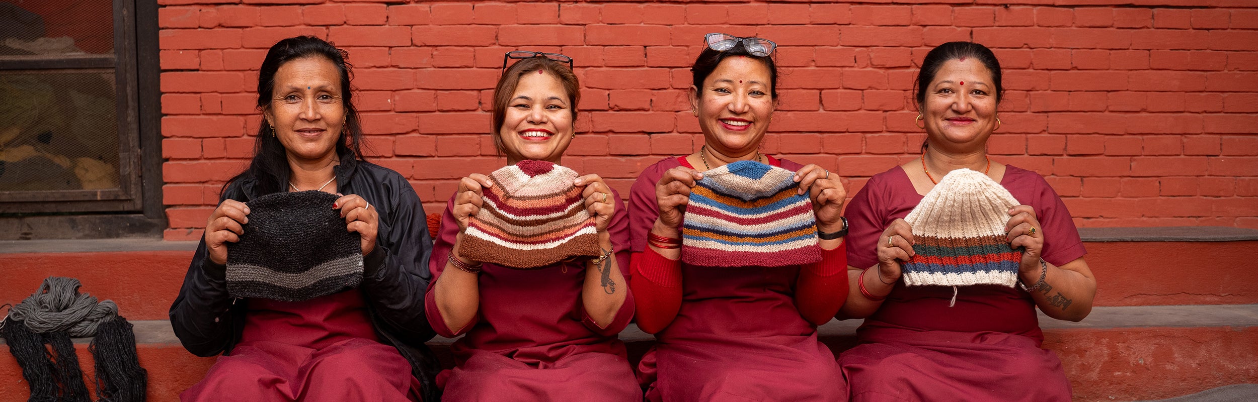 A group of women in Nepal, sitting in a line on a set of steps. They are smiling at the camera holding up hats they have knitted.