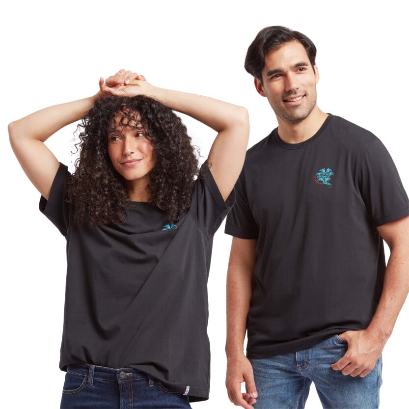 A man and a woman wearing Sherpa Adventure Gear Deity T-Shirt in Black. The man’s T-shirt fits snugly, paired with blue jeans. The woman wears a slightly oversized fit, also paired with jeans. Both shirts display a small turquoise and red lotus flower design on the chest. The background is a clean, white studio setup.