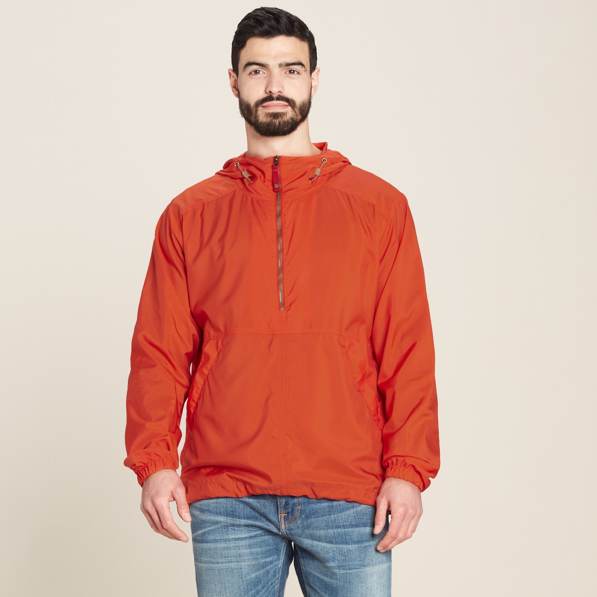 A man with a beard and short dark hair is wearing a Sherpa Adventure Gear Halka Anorak Jacket in Red, standing against a neutral beige background. The jacket has a front half-zip closure, elastic cuffs, and a kangaroo pocket. The model looks directly into the camera with a neutral expression, his arms relaxed at his sides, showcasing the jacket's relaxed fit.