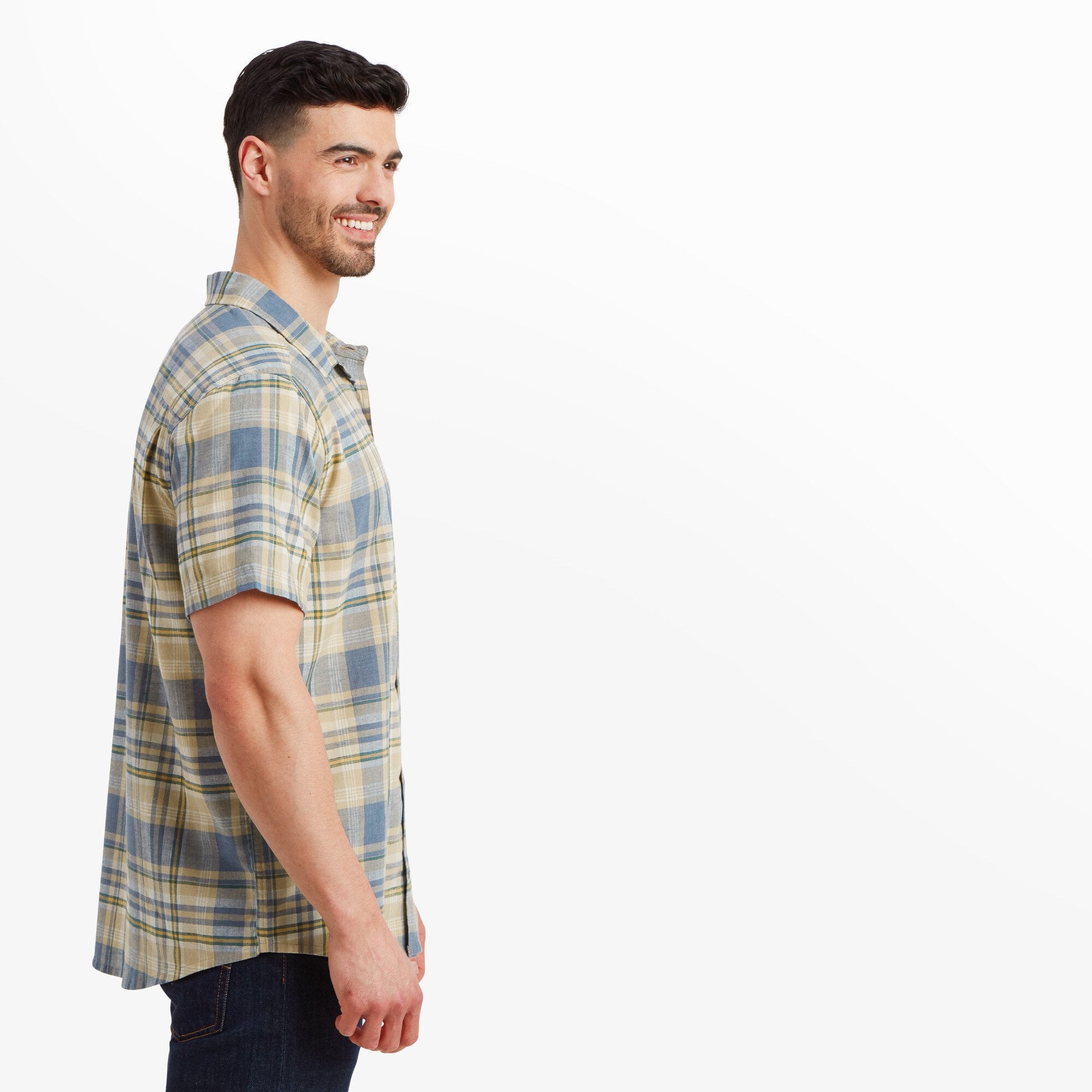 A side profile of the same man wearing the Sherpa Adventure Gear Hosh Plaid Short Sleeve Shirt in Beige. The image highlights the shirt’s tailored fit and short sleeve length, showing how it falls naturally on the body. The model is smiling and looking forward.