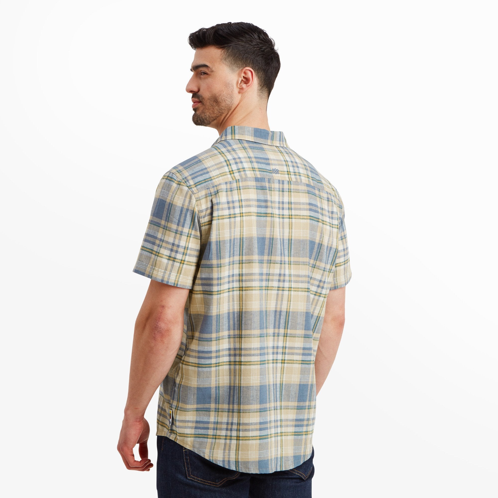 The man facing away from the camera, displaying the back of the Sherpa Adventure Gear Hosh Plaid Short Sleeve Shirt in Beige. The pattern continuity and clean cut of the shirt are visible, showcasing its design from behind.