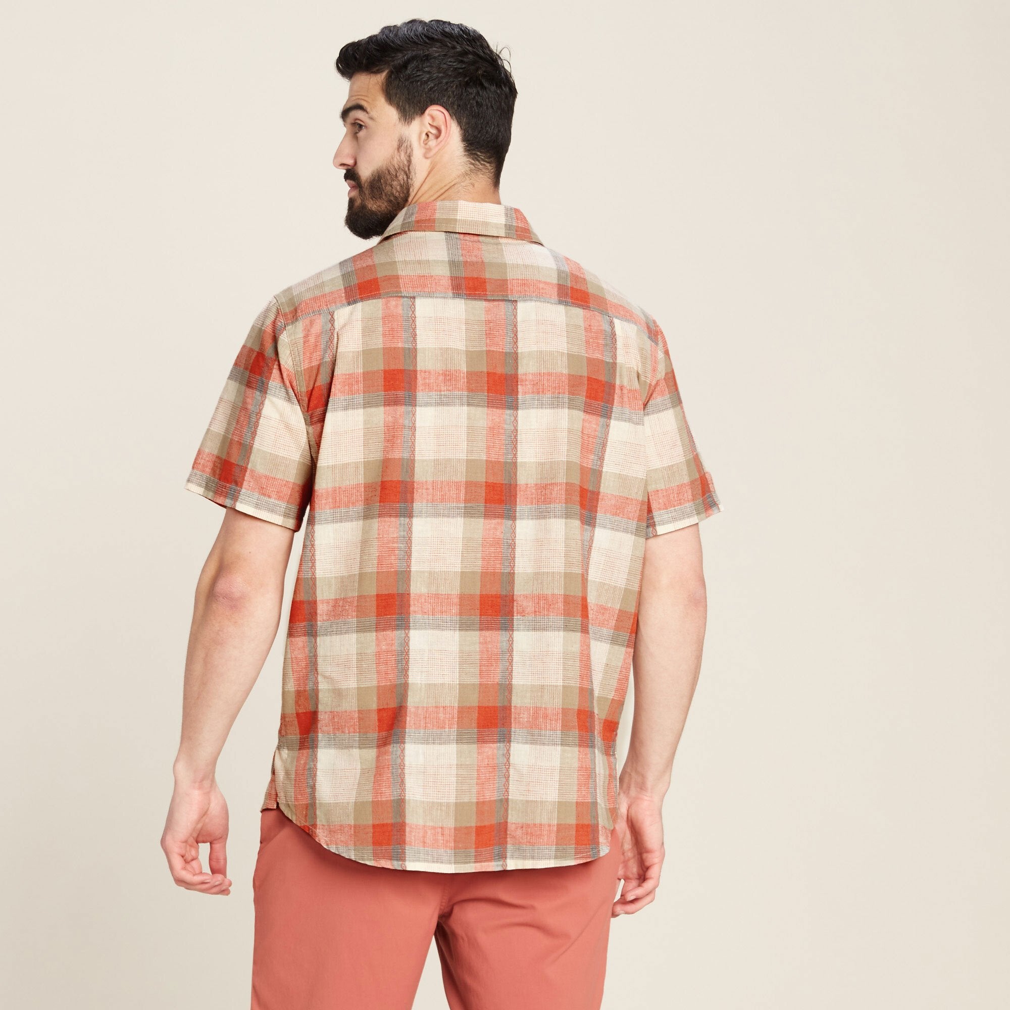 The back of the Sherpa Adventure Gear Khamsu Short Sleeve Shirt in Grey showcases its evenly spaced checkered pattern in earthy tones. The model is turned away, highlighting the shirt's straight hemline and comfortable fit.