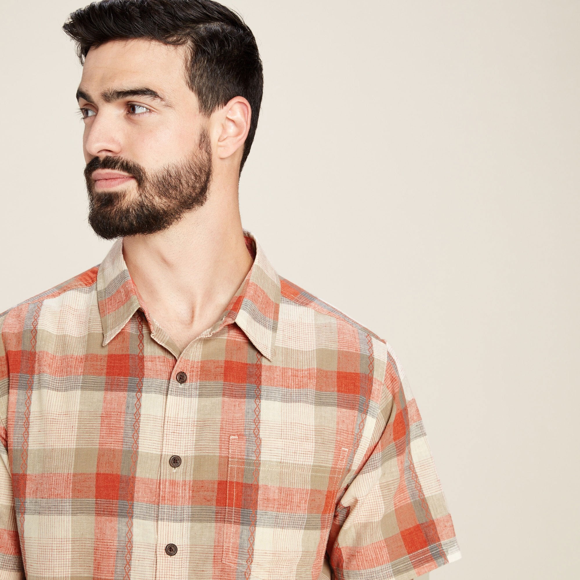 A closer view of the Sherpa Adventure Gear Khamsu Short Sleeve Shirt in Grey emphasises the intricate stitching and soft texture of the fabric. The subtle mix of red and beige tones adds warmth, while the collar and buttons give the shirt a polished, yet casual appearance.