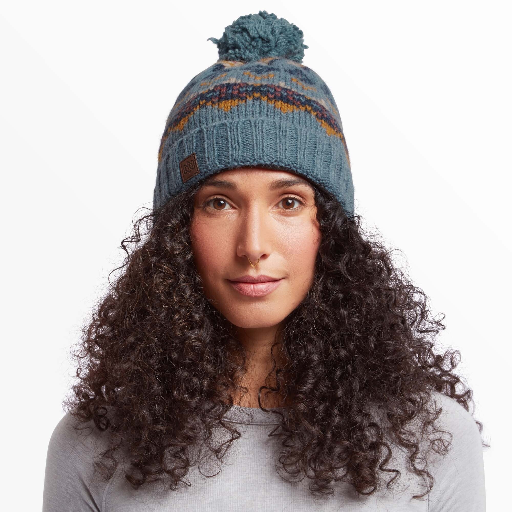 A close-up view of a woman wearing a Sherpa Adventure Gear Lahan Hat in Blue with a pom-pom on top. The beanie features a zigzag multicolor pattern in warm tones, such as orange and brown, and a brown leather patch with a logo on the front. The woman has curly dark hair cascading around the hat and is wearing a light gray shirt, with a serene expression on her face.