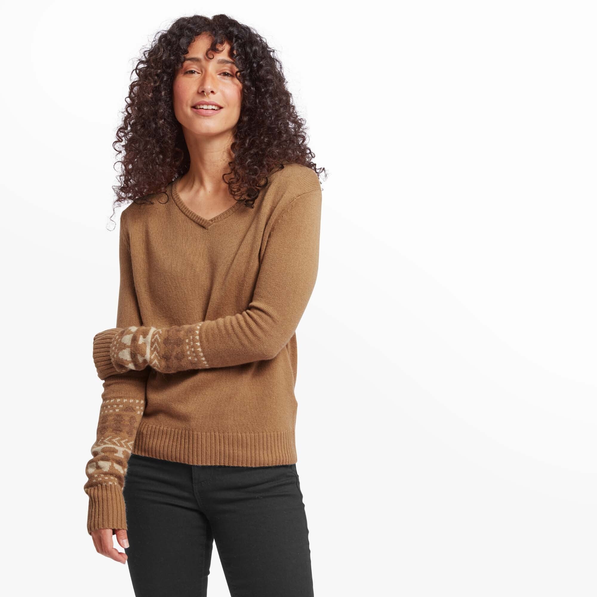 The first image features a woman with curly hair wearing a Sherpa Adventure Gear Maya V-Neck Sweater in Light Brown. The sweater has intricate beige and white patterns along the forearms. She is standing with one arm across her torso, showcasing the patterned details of the sleeves. Her black jeans and the sweater's casual style create a warm and relaxed appearance.