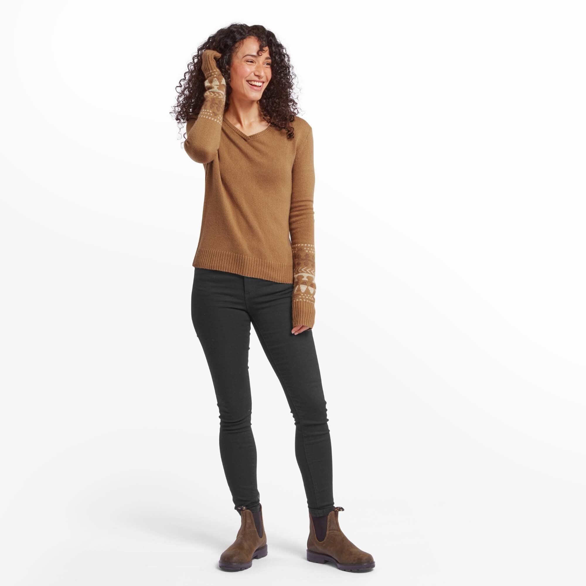 The second image depicts the same woman standing fully, wearing the Sherpa Adventure Gear Maya V-Neck Sweater in Light Brown paired with black skinny jeans and brown ankle boots. She is smiling and holding her curly hair back, exuding confidence and a cozy yet chic style.