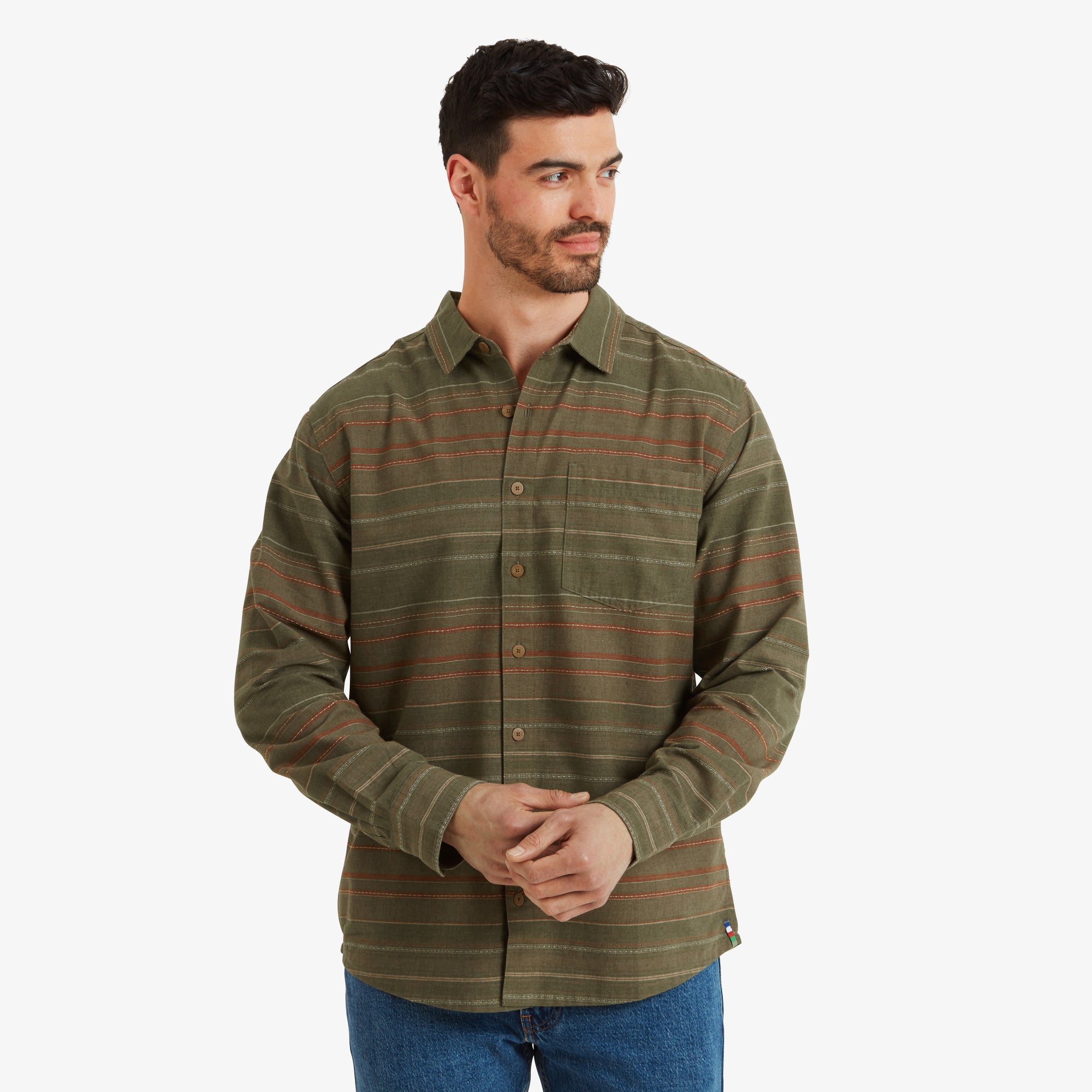 A man wearing a Sherpa Adventure Gear Sangpo Long Sleeve Shirt in Grey with subtle horizontal stripes in earthy tones. He is standing with hands clasped and gazing slightly to the side. The shirt features a single chest pocket, and the model pairs it with light blue denim jeans.