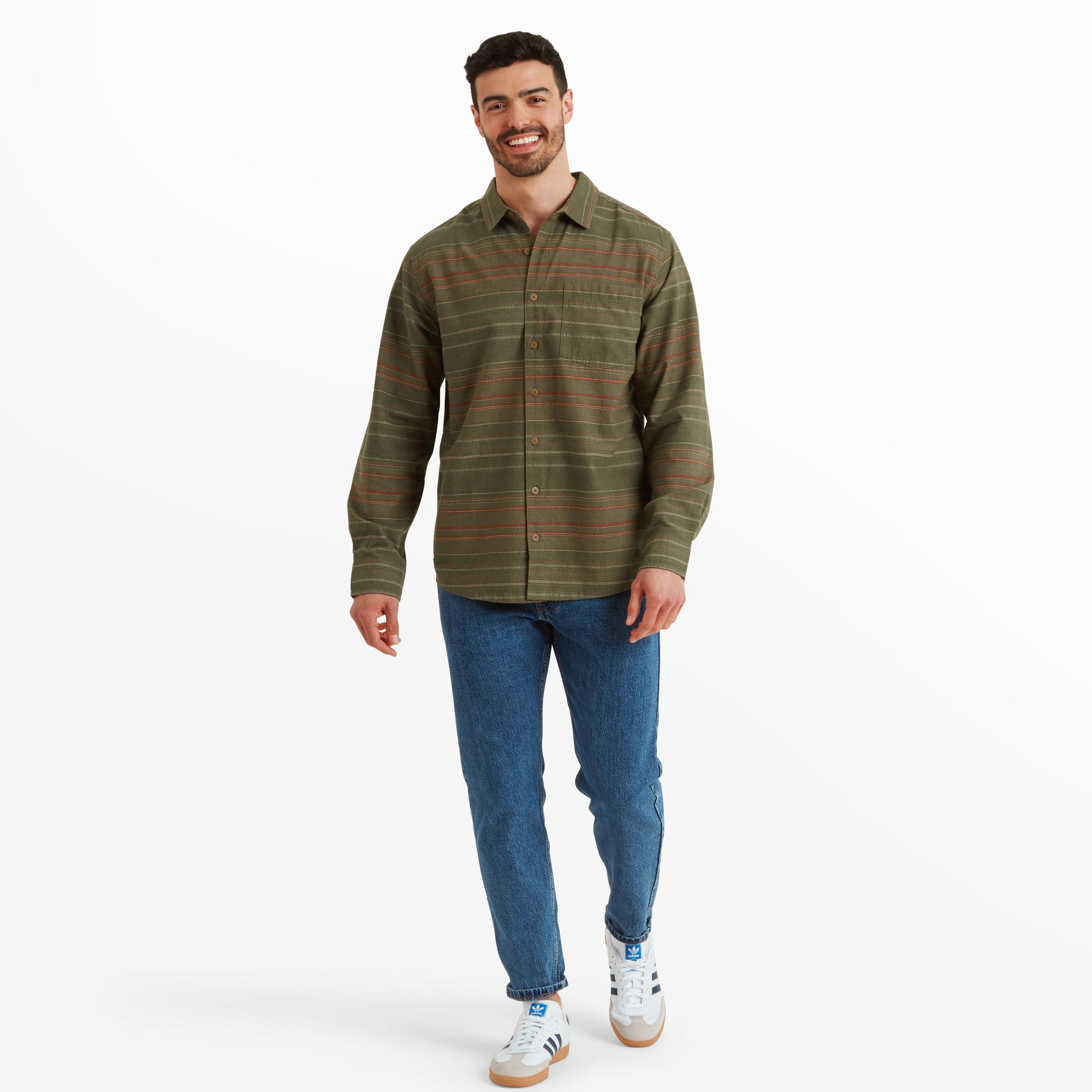 The same man is shown in a full-body shot walking forward while smiling. He is dressed in the Sherpa Adventure Gear Sangpo Long Sleeve Shirt in Grey, tucked out over light blue denim jeans, complemented by casual white trainers with blue accents.