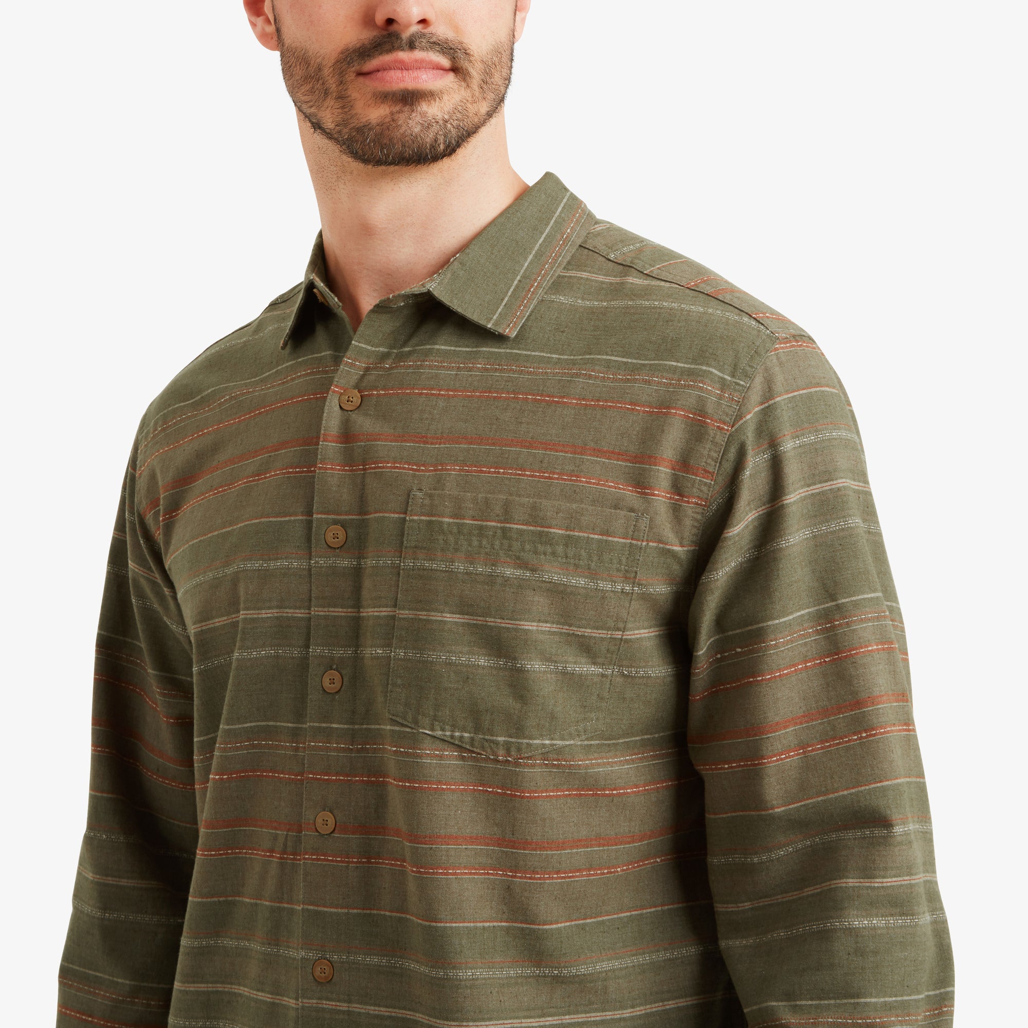 A close-up of the upper torso of the model wearing the Sherpa Adventure Gear Sangpo Long Sleeve Shirt in Grey. The details of the fabric, button closures, and chest pocket are visible, emphasising the subtle contrast of the earthy-toned stripes. The model gazes slightly to the side.