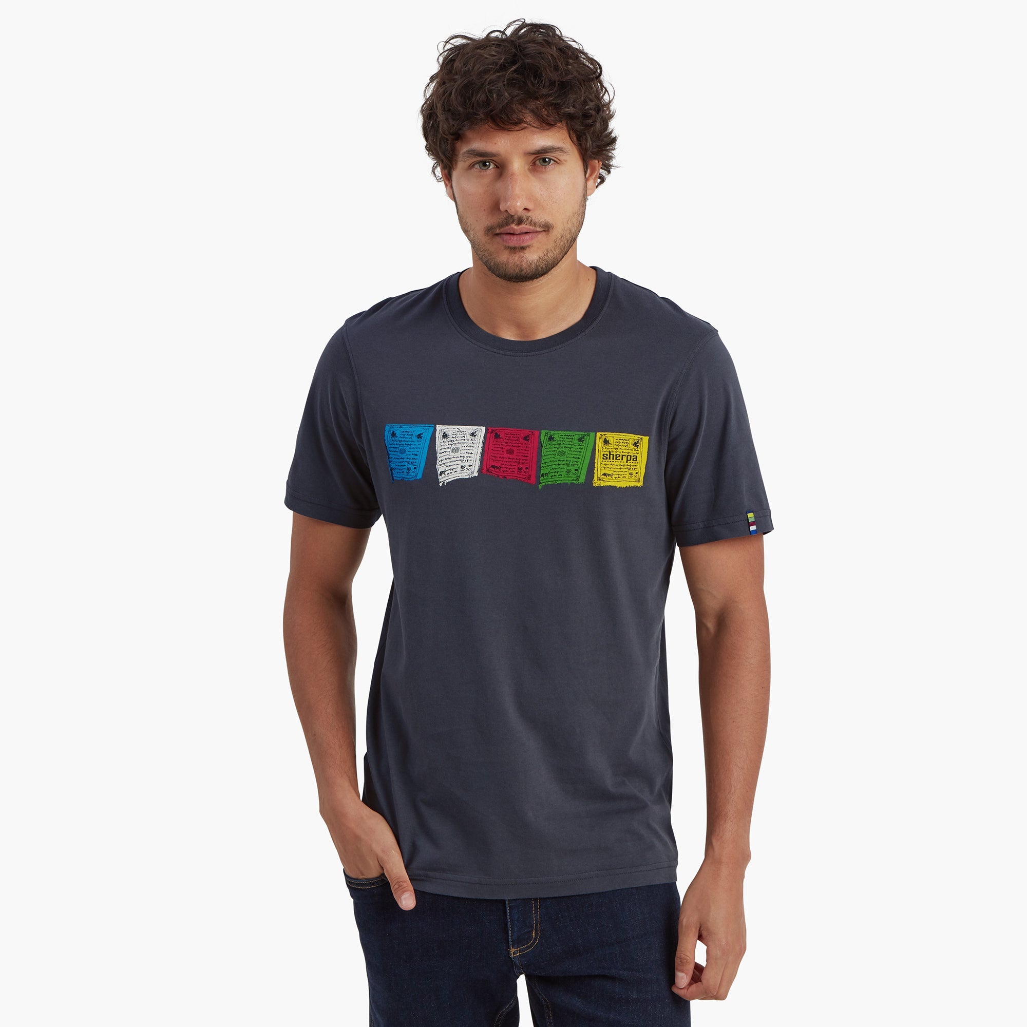 A male model wearing a Sherpa Adventure Gear Tarcho T-Shirt in Grey featuring colorful Tibetan prayer flags printed across the chest in blue, white, red, green, and yellow. He is standing straight with one hand in his pocket, showcasing the shirt's fit and design.