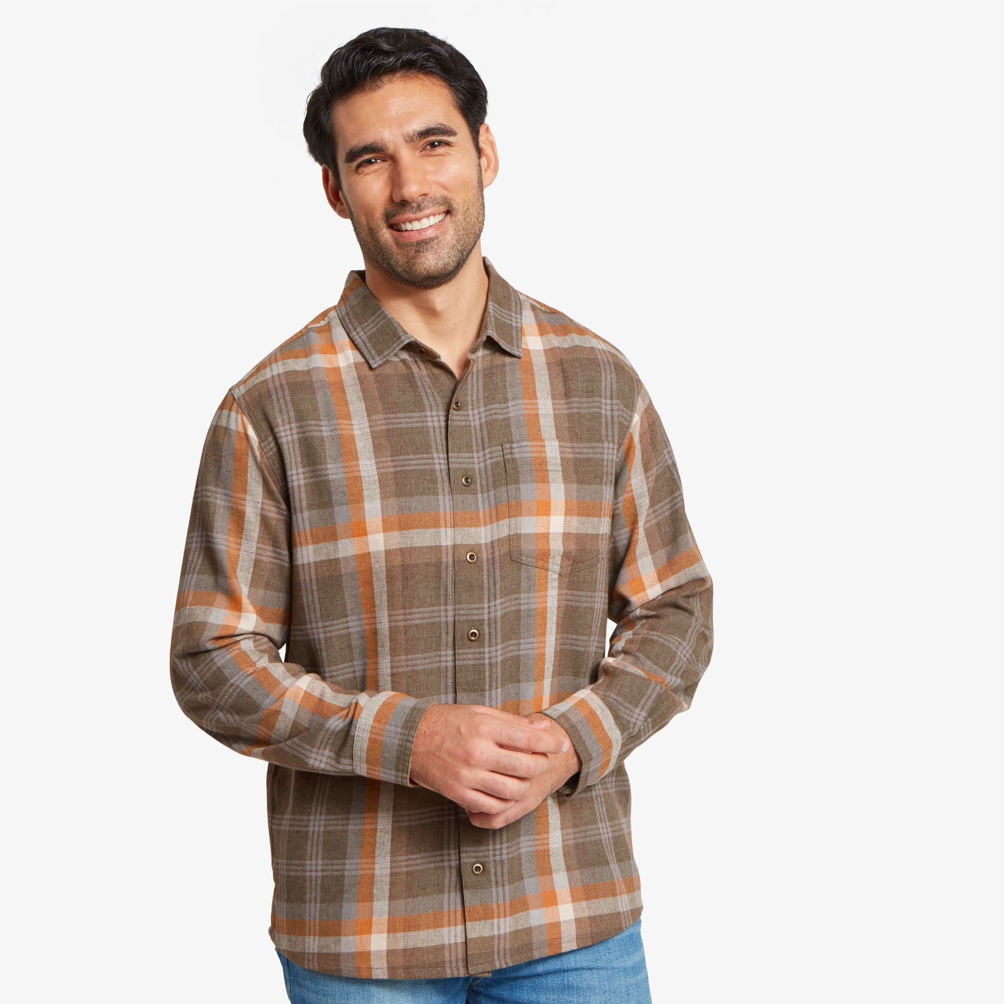 A front-facing shot of a man wearing a Sherpa Adventure Gear Yama Long Sleeve Shirt in Beige. He is smiling and has his hands clasped in front of him. The shirt features a classic button-up design with a left chest pocket, and the model is wearing light blue jeans.