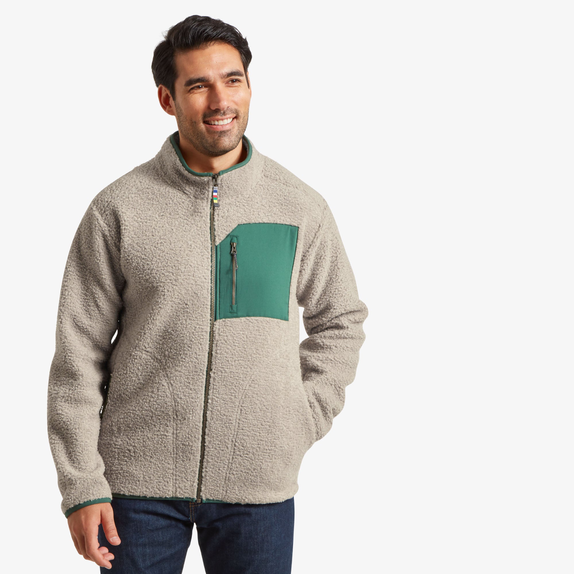 A man is wearing a Sherpa Adventure Gear Yatri Full Zip Jacket in Beige with a green chest pocket featuring a zippered closure. The jacket has a cozy texture and a high collar, paired with blue denim jeans. The front view highlights the sleek, functional design and warm material.