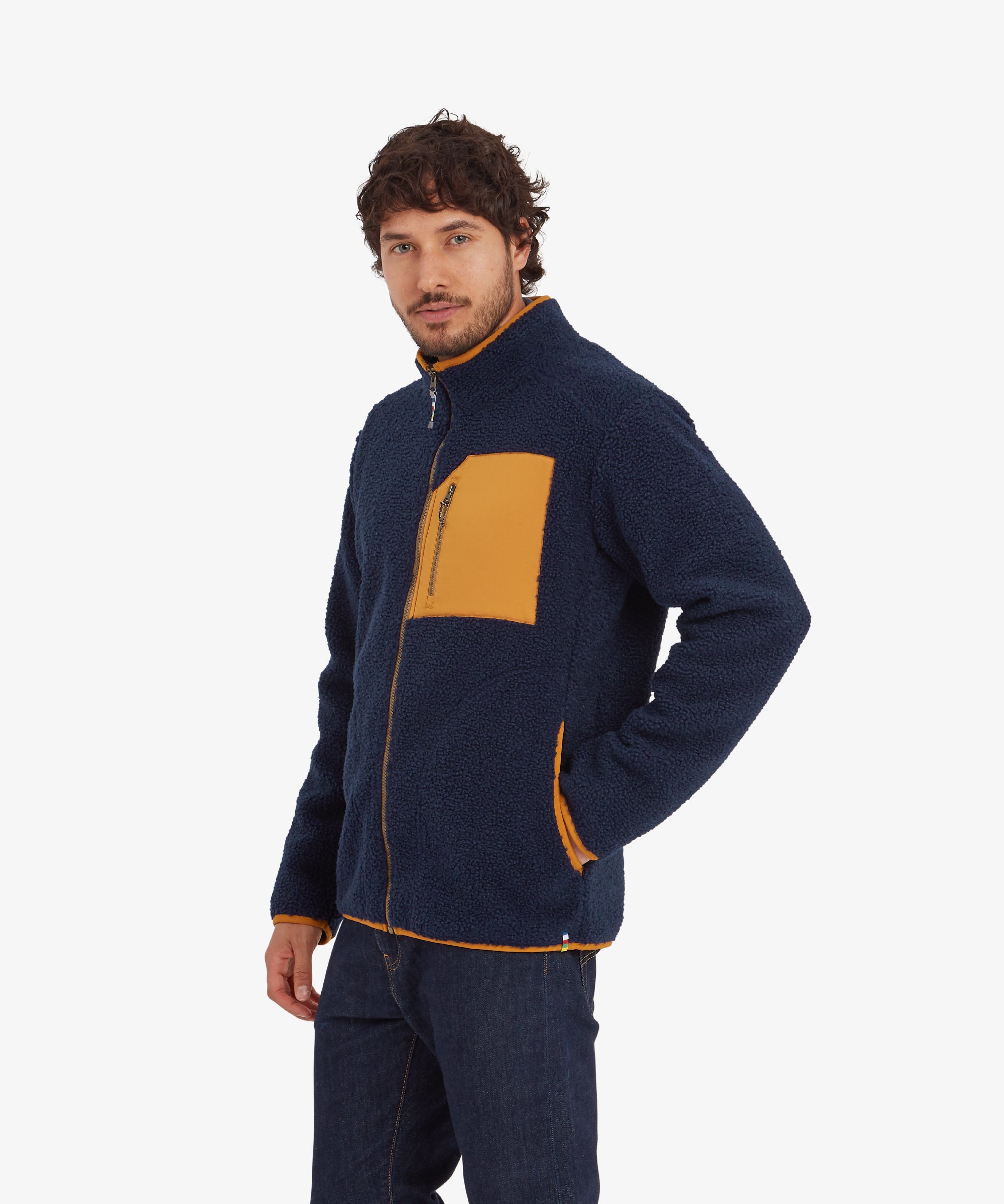 A side profile view of the Sherpa Adventure Gear Yatri Full Zip Jacket in Blue, highlighting the zippered mustard chest pocket and orange edge piping. The man looks to the side, showcasing the jacket's structured fit and side pocket detail.