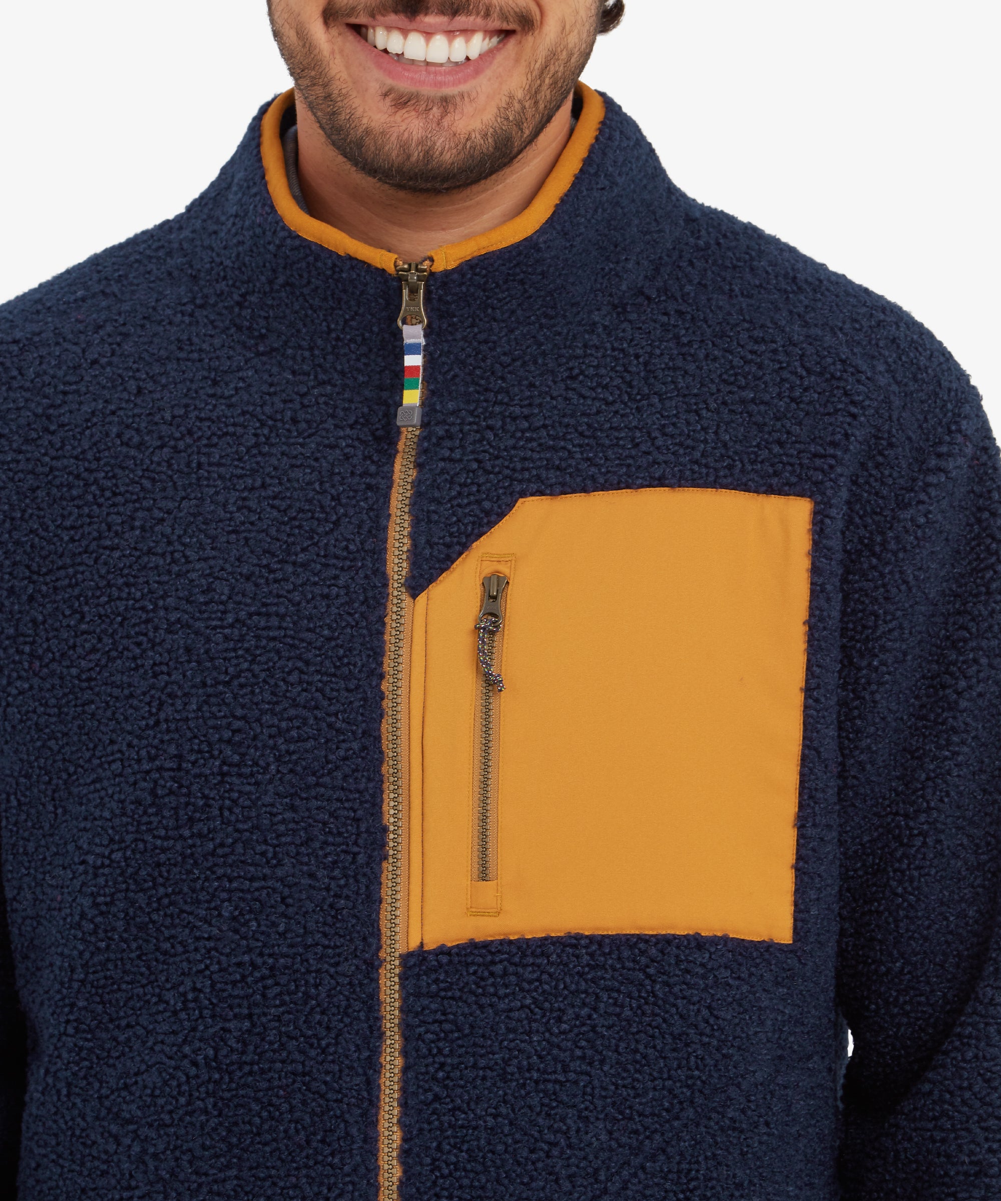 A close-up of the mustard yellow zippered chest pocket on the Sherpa Adventure Gear Yatri Full Zip Jacket in Blue. The zipper pull has a braided detail, and the contrasting textures between the fleece and pocket material are clearly visible.