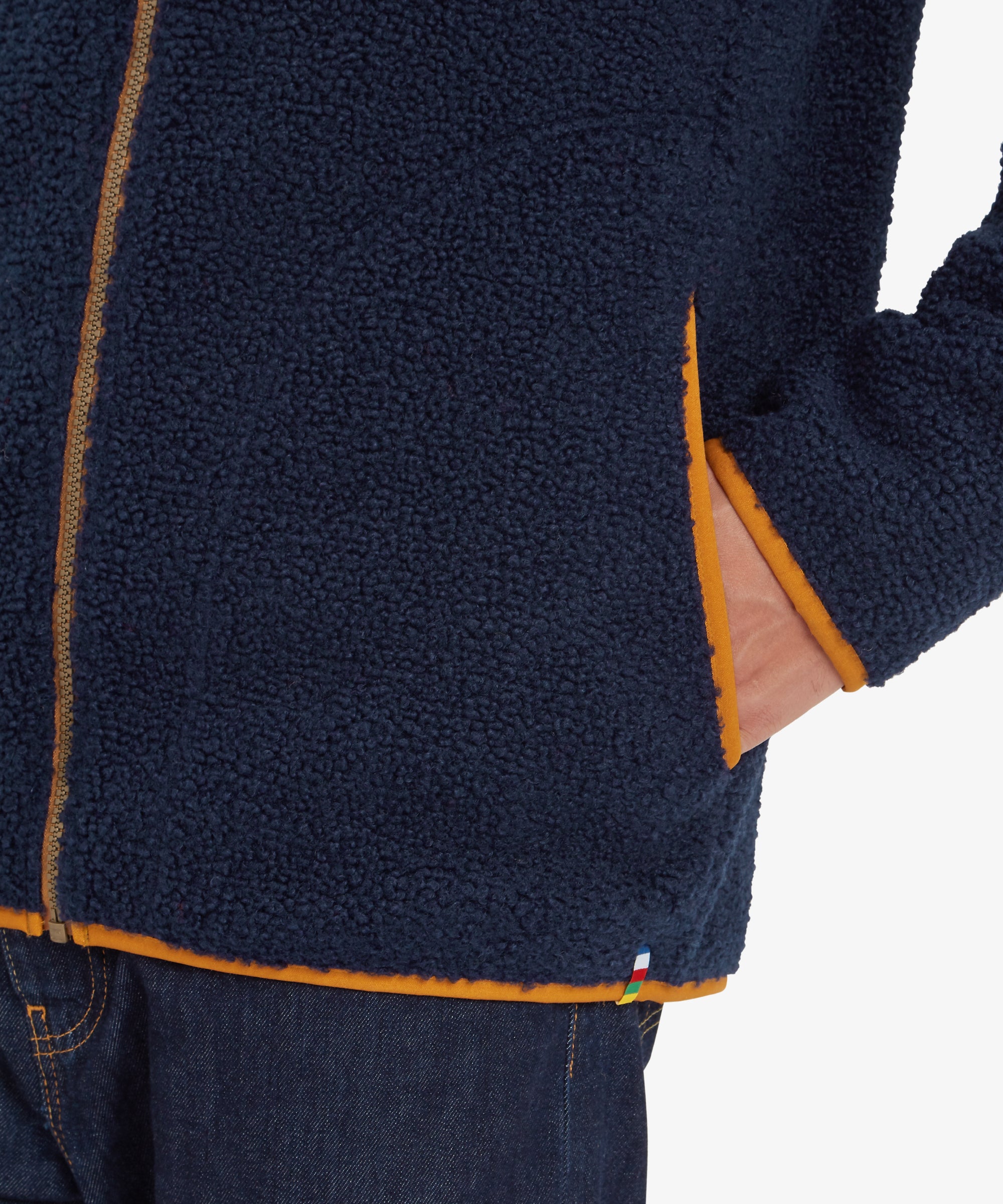 A zoomed-in view of the lower edge of the Sherpa Adventure Gear Yatri Full Zip Jacket in Blue. The side pocket opening and orange piping are visible, along with a small multicolor striped label near the hem, emphasising the garment's craftsmanship.