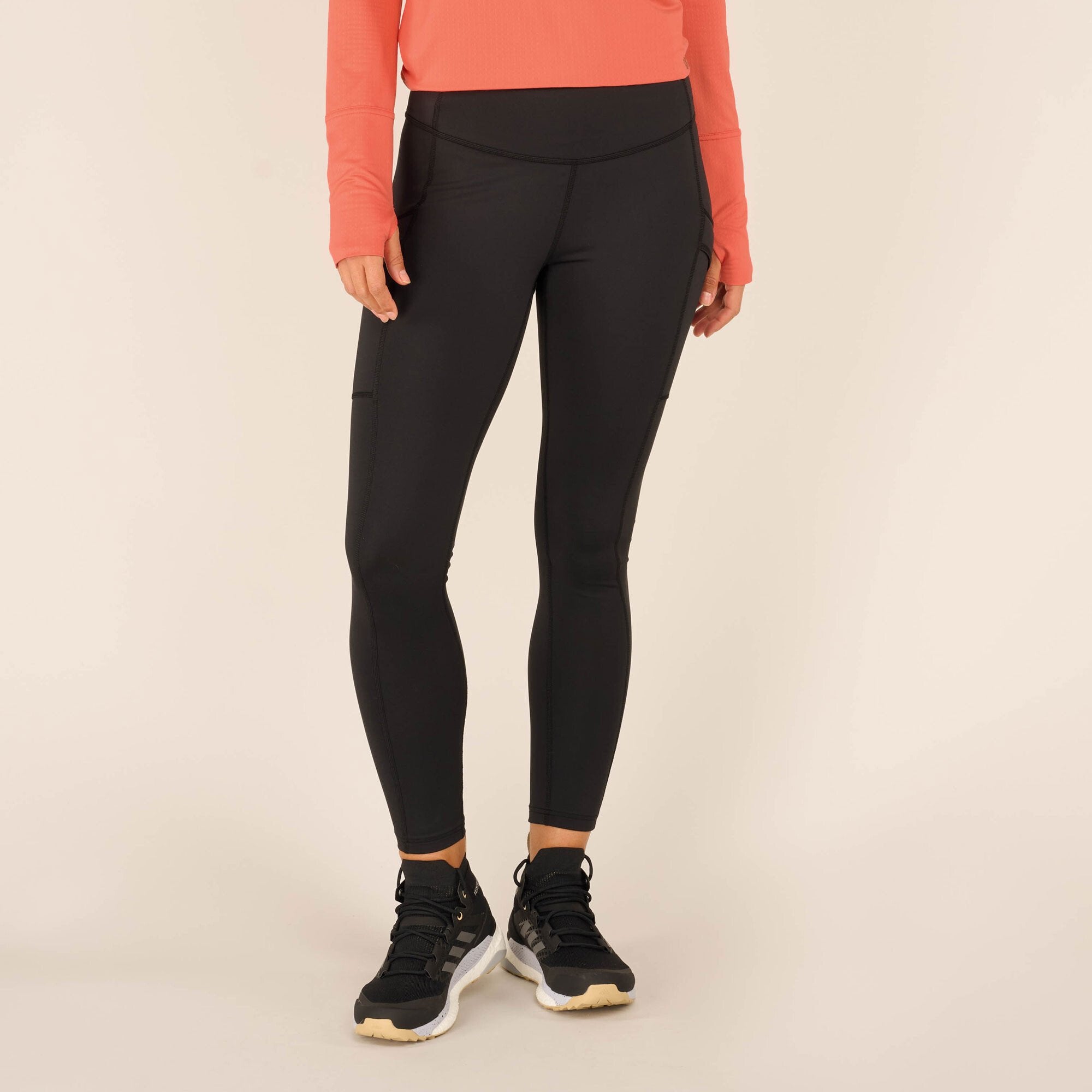 A close-up view of a model’s lower body, showcasing Sherpa Adventure Gear Nisha Tight in Black. The leggings fit snugly, contouring the model’s legs and waist. The fabric appears smooth and stretchy, with visible seams running along the sides and around the hips. The model is wearing black trainers with a chunky white sole.