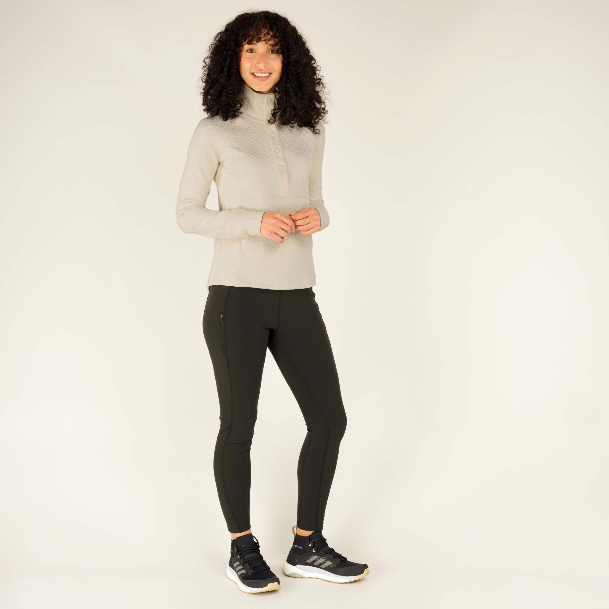 The woman is seen standing in a casual pose, hands lightly clasped, while wearing the Sherpa Adventure Gear Nyano Snap Mock Neck in Green and black leggings. The full-length view emphasises the pullover’s comfortable fit and stylish design. She complements the outfit with black trainers featuring white soles.