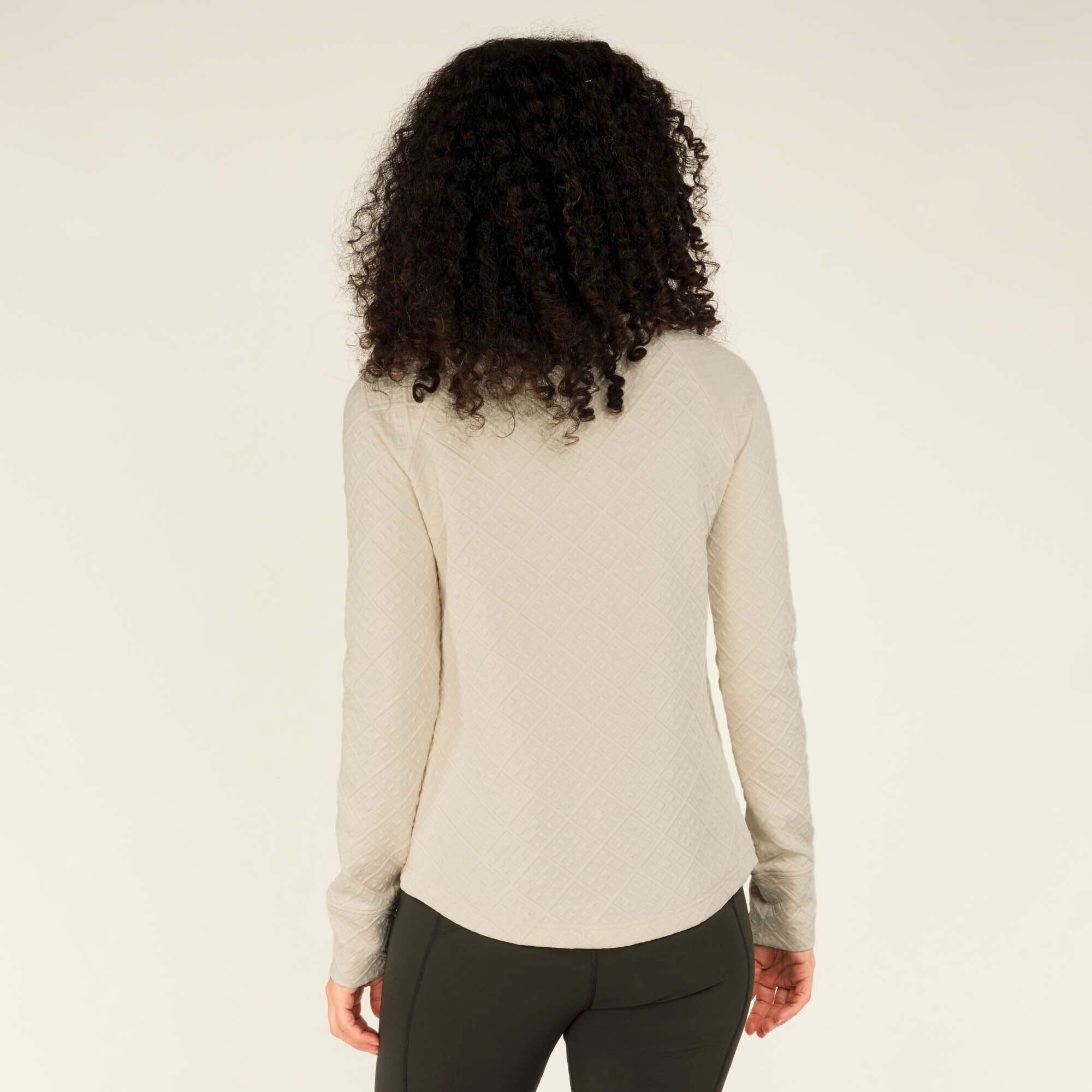 The image highlights the back view of the Sherpa Adventure Gear Nyano Snap Mock Neck in Green, displaying the subtle curvature of the hemline and the full quilted texture. The black leggings create a sleek contrast with the lighter tone of the top, and her curly hair adds a playful, natural element to the overall look.