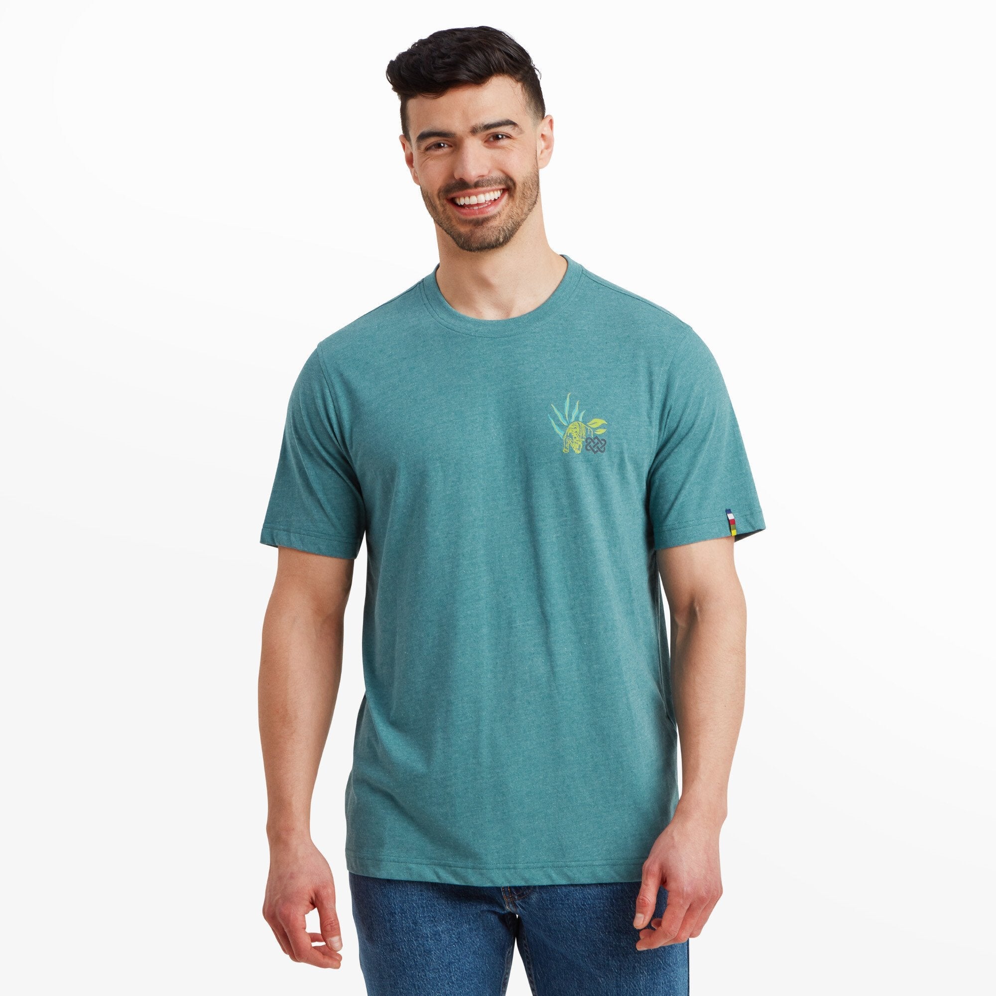 A man is wearing a Sherpa Adventure Gear Purpose T-Shirt in Blue with a small graphic on the chest, featuring a yellow tiger surrounded by stylized green leaves. The T-shirt has a classic crew neck and fits comfortably. The man is smiling, and the background is plain white, drawing attention to the shirt.