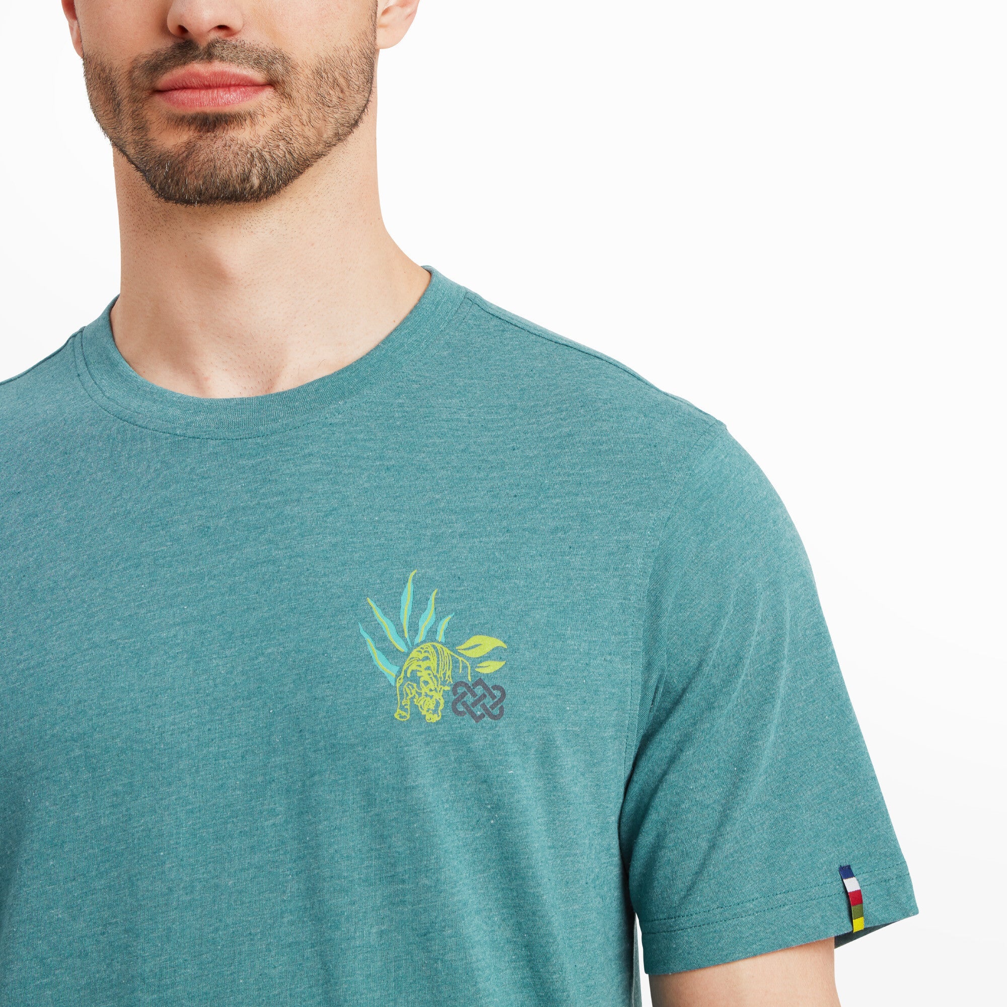 A zoomed-in shot of the chest graphic focuses on the small design of the tiger and leaves. The intricate details in the artwork and the subtle placement on the Sherpa Adventure Gear Purpose T-Shirt in Blue add an element of sophistication and style to the piece. The multicolored Sherpa logo flag on the sleeve is also visible, complementing the overall design.