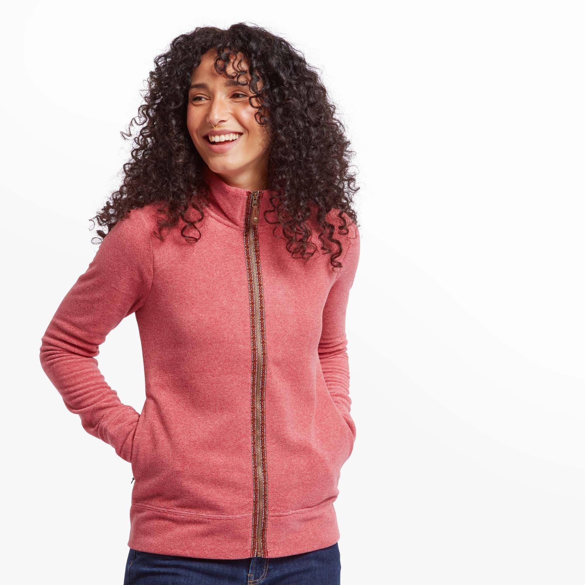 A smiling woman wearing a Sherpa Adventure Gear Rolpa Eco Jacket in Red. The jacket features a full front zipper with a patterned trim and a high collar. She has her hands in the jacket pockets and is standing against a plain white background.