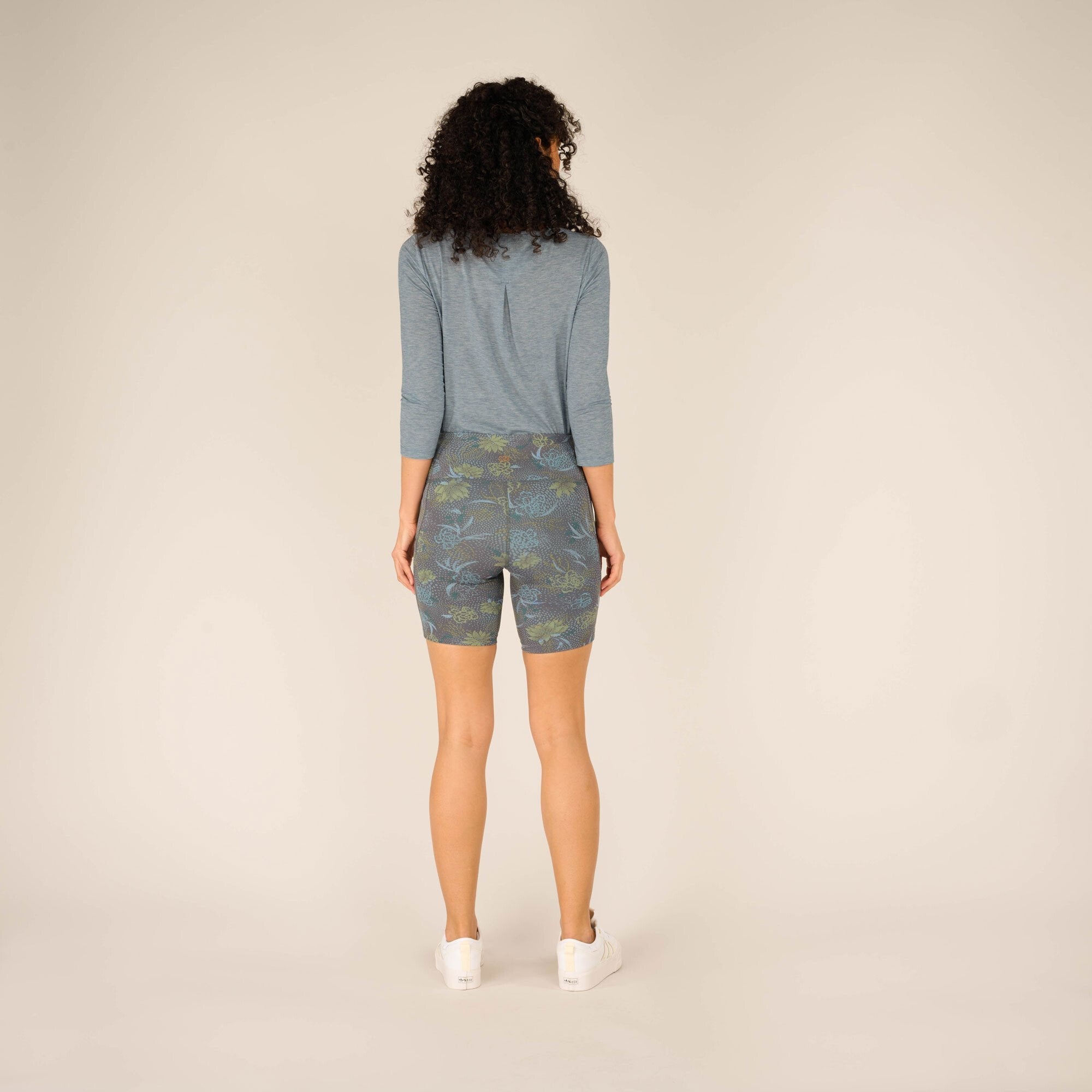 A complete view of the model standing with their back turned, showcasing the back fit of the Sherpa Adventure Gear Sahasa Shorts in Blue. The light blue top and trainers add balance to the vibrant print of the shorts.
