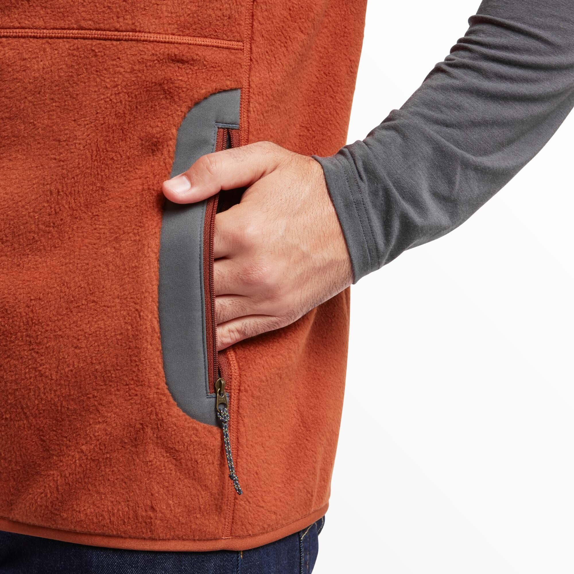 Close-up of a person wearing a Sherpa Adventure Gear Sanani Eco Fleece Vest in Red, showcasing the side pocket detail. The pocket is outlined with a contrasting gray trim and features a zippered closure with a decorative pull tab. The person's hand is partially inserted into the pocket, emphasising its size and functionality. The texture of the fleece material is visible, highlighting its soft and cozy appearance. A gray long-sleeve shirt is worn underneath, complementing the gray accents on the vest.