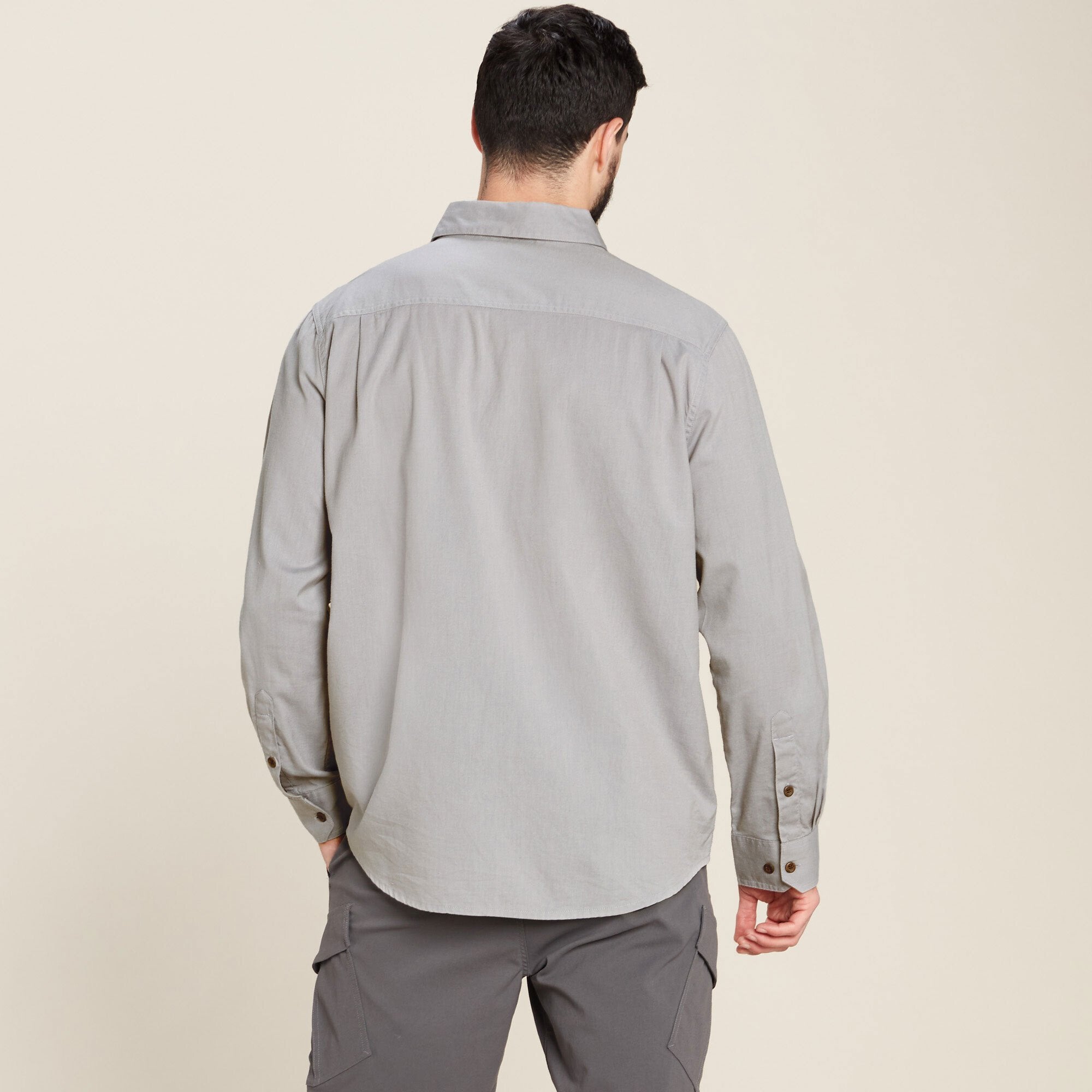 The back view of the same Sherpa Adventure Gear Tharu Long Sleeve Shirt in Grey, showcasing its clean and minimalist design. The model has his arms relaxed at his sides, highlighting the back yoke and curved hemline of the shirt.