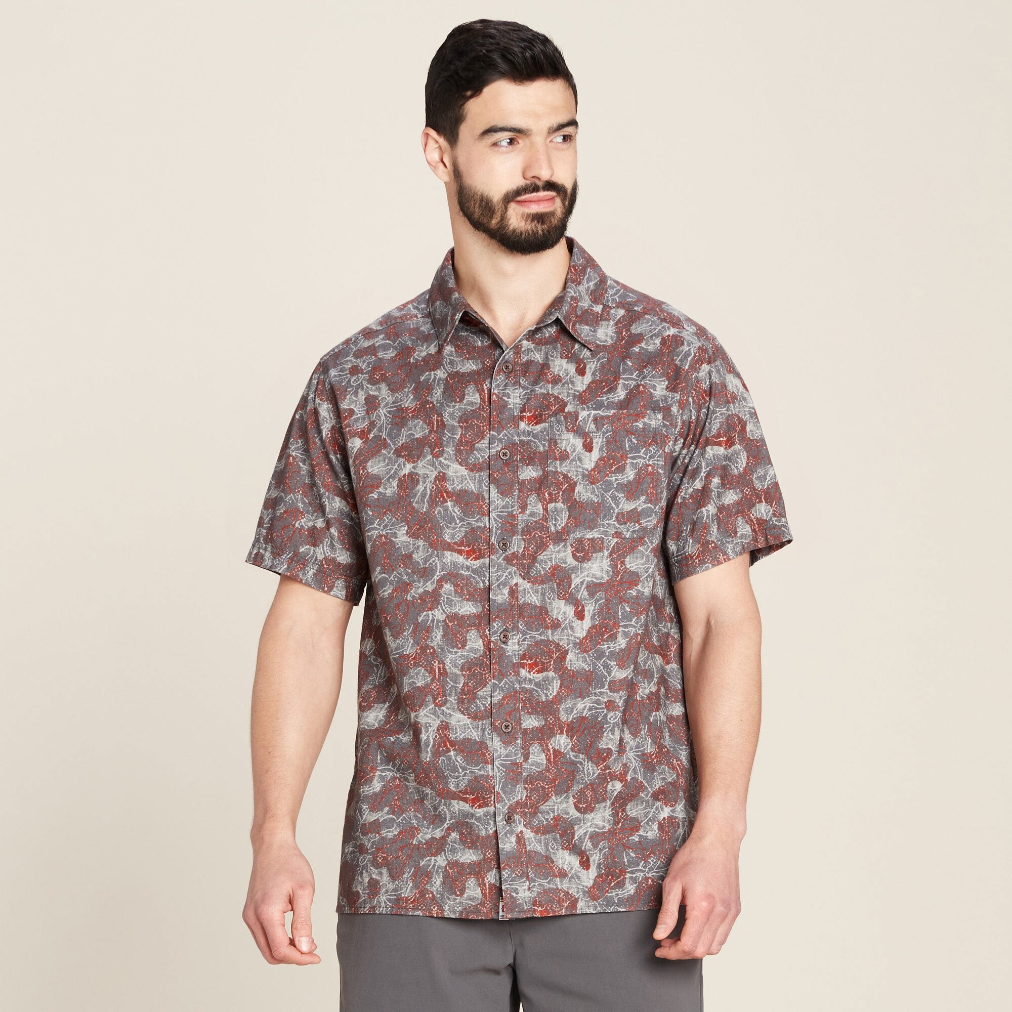 A man modeling a Sherpa Adventure Gear Tharu Voyage Short Sleeve Shirt in Grey featuring a unique abstract pattern in shades of grey and red. The shirt has a relaxed fit and a spread collar, with buttons running down the front. The model is styled with a neutral beige background, showcasing the garment's casual and versatile design.