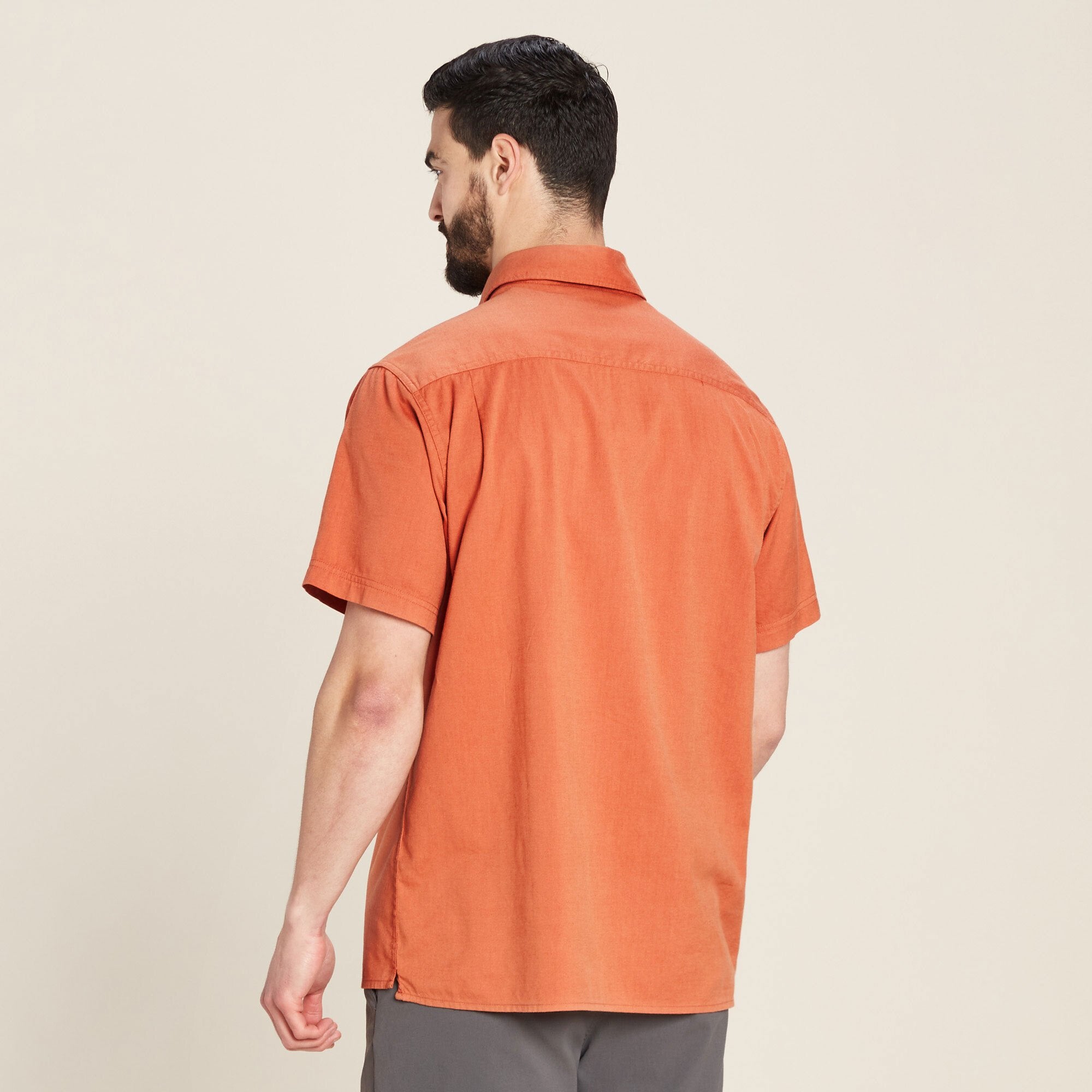 The model is turned away, showing the back of the Sherpa Adventure Gear Tharu Voyage Short Sleeve Shirt in Orange. The back highlights a clean and seamless design without extra embellishments, emphasising the simplicity and versatility of the shirt.