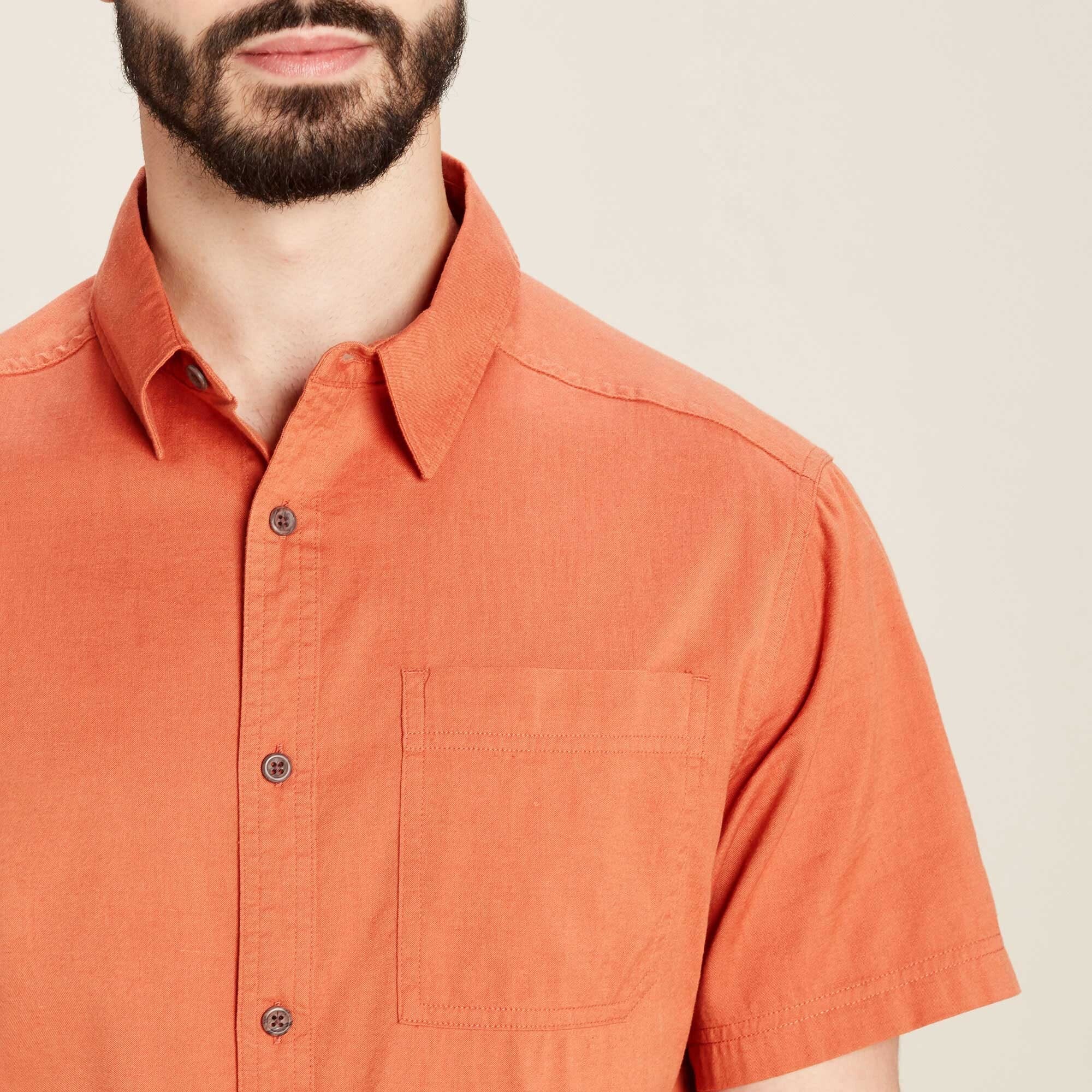 A detailed close-up of the Sherpa Adventure Gear Tharu Voyage Short Sleeve Shirt in Orange's front upper section, focusing on the neatly sewn front pocket, the buttons, and the crisp collar. The warm orange fabric is slightly textured, suggesting durability and comfort, making it suitable for casual outings or light travel.