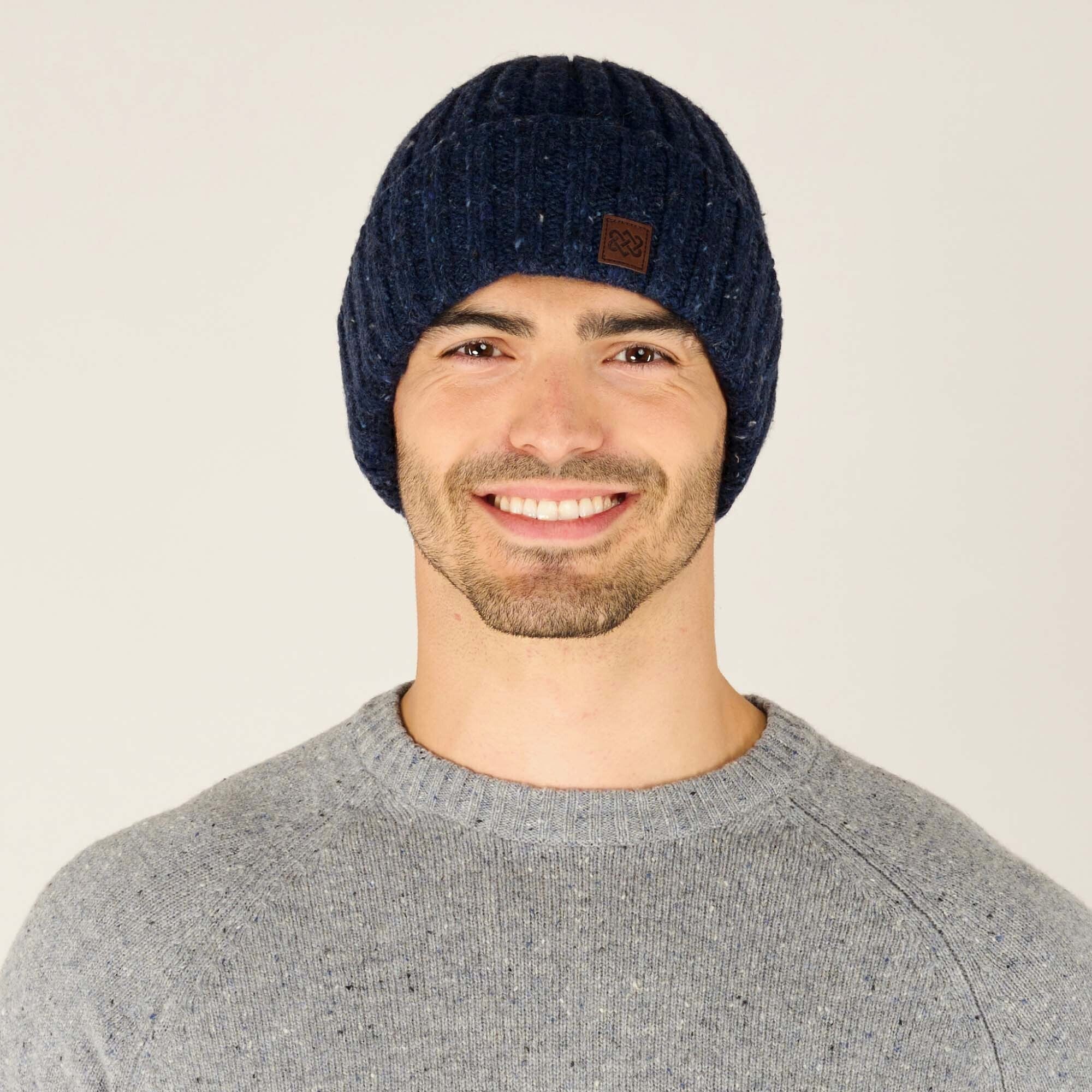 A close-up shot of a smiling model wearing a Sherpa Adventure Gear Varuni Classic Beanie in Blue with speckled white and lighter blue details. The beanie features a small brown leather patch with an embossed knot design on the front. The model wears a light grey textured sweater, complementing the beanie’s understated yet stylish appearance.