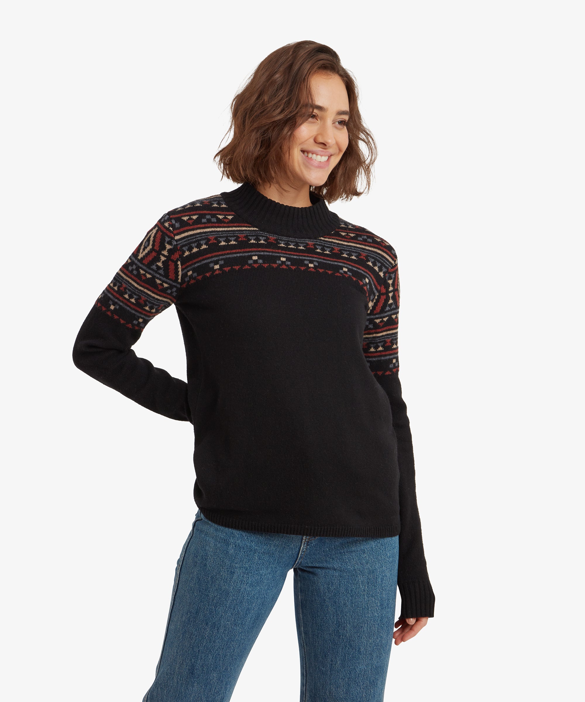 A woman with shoulder-length brown hair and a smile is wearing a Sherpa Adventure Gear Bhutla Crew Sweater in Black  with intricate red, beige, and blue geometric patterns across the chest and upper arms. She stands slightly turned, with one hand behind her back and the other relaxed. The sweater has a ribbed crew neck and long sleeves with ribbed cuffs.