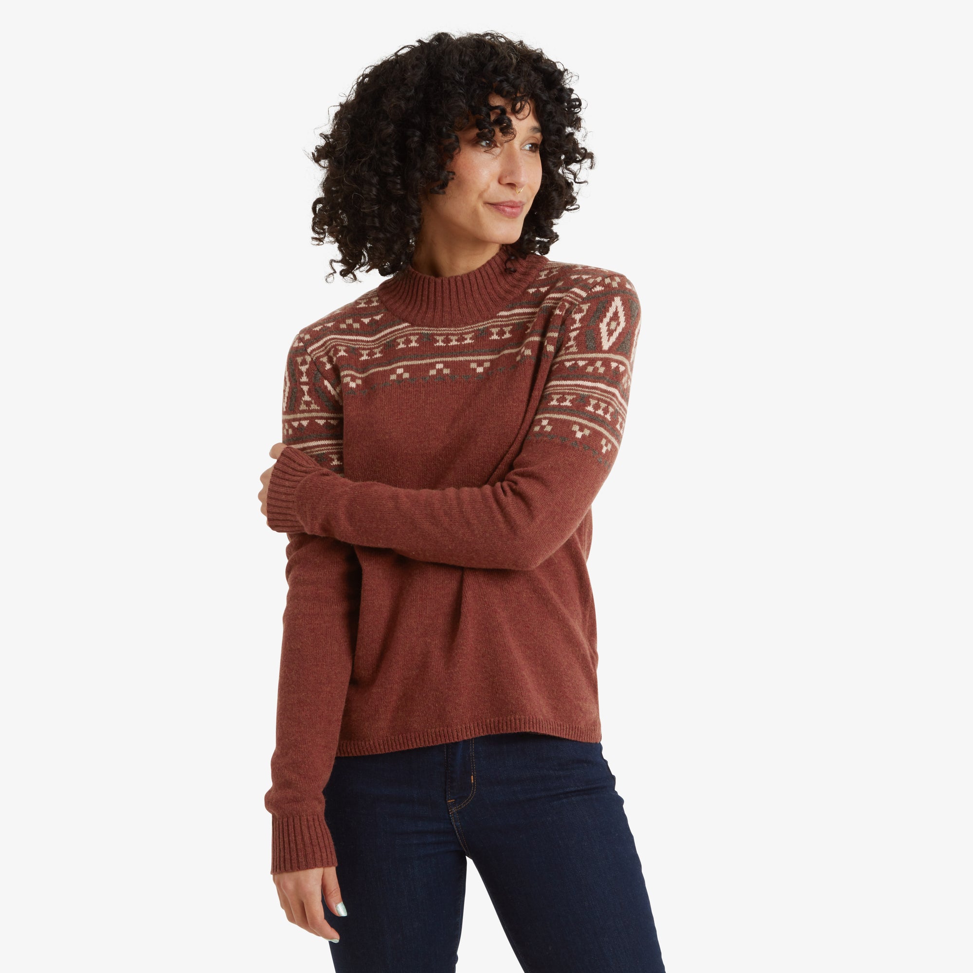 A woman is seen wearing a Sherpa Adventure Gear Bhutla Crew Sweater in Brown featuring traditional geometric patterns in beige, white, and green across the chest, shoulders, and upper sleeves. The sweater has a high ribbed crew neckline and long ribbed cuffs. She pairs the sweater with dark blue jeans. Her curly black hair frames her face as she looks to her left with a soft smile. The setting is a clean white background.