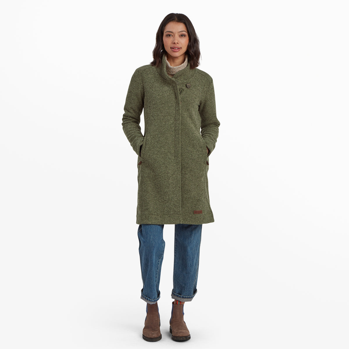 Eine Frau steht mit Blick nach vorne und trŠgt eine Sherpa Adventure Gear Lhamu Jacke in GrŸn mit hohem Kragen und einem einzelnen Knopfdetail. Der Mantel hat zwei Seitentaschen und wird mit einer Jeans mit Umschlag und braunen Stiefeletten mit blauen Akzenten kombiniert. Ihre HŠnde ruhen entspannt in den Manteltaschen und sie hat einen neutralen Gesichtsausdruck.