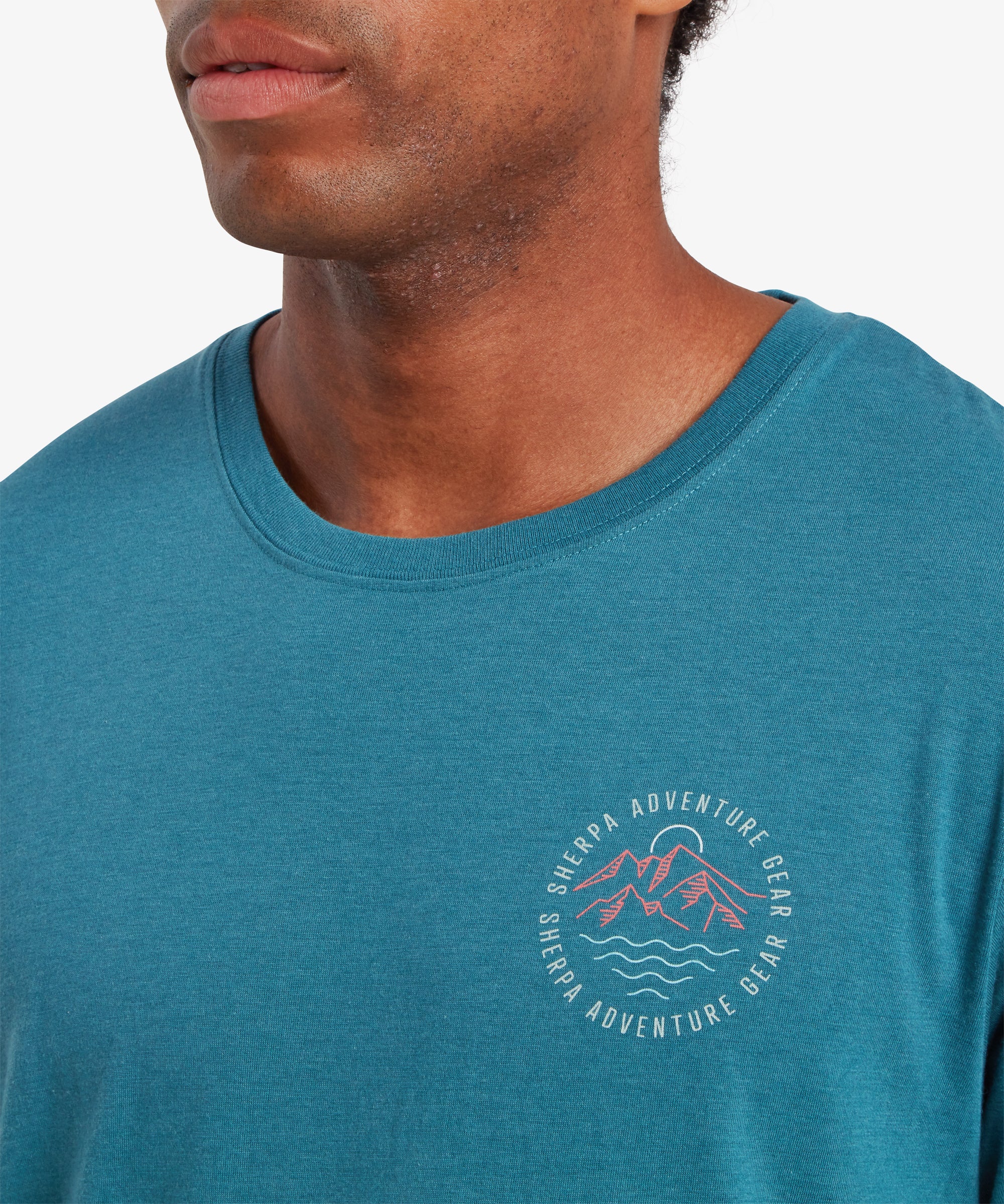 Close up of a man wearing a blue t-shirt with a printed logo on the left chest. The logo depicts a graphic mountain range in red with a white river below and a white sun above. The words "Sherpa Adventure Gear" is written around the logo in a circle.